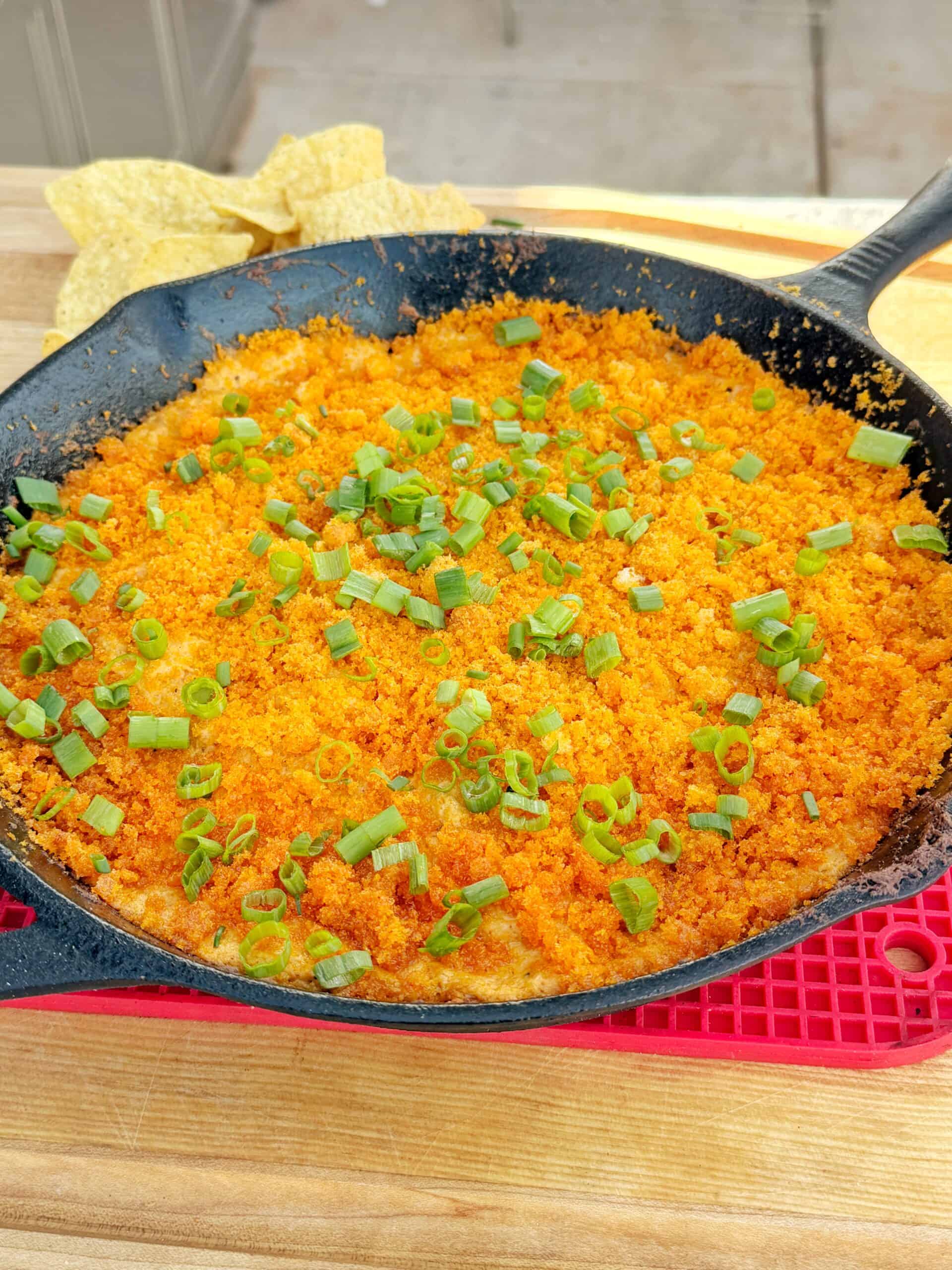 A cast iron skillet filled with a golden, crispy baked dish topped with chopped green onions, resting on a red trivet. Tortilla chips are visible in the background on a wooden surface.