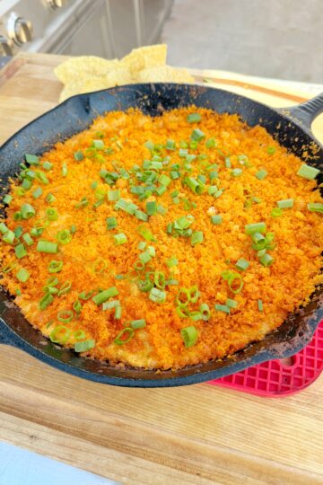 A cast iron skillet filled with a baked dish topped with orange breadcrumbs and chopped green onions, sitting on a wooden surface with tortilla chips and a red hot pad nearby.