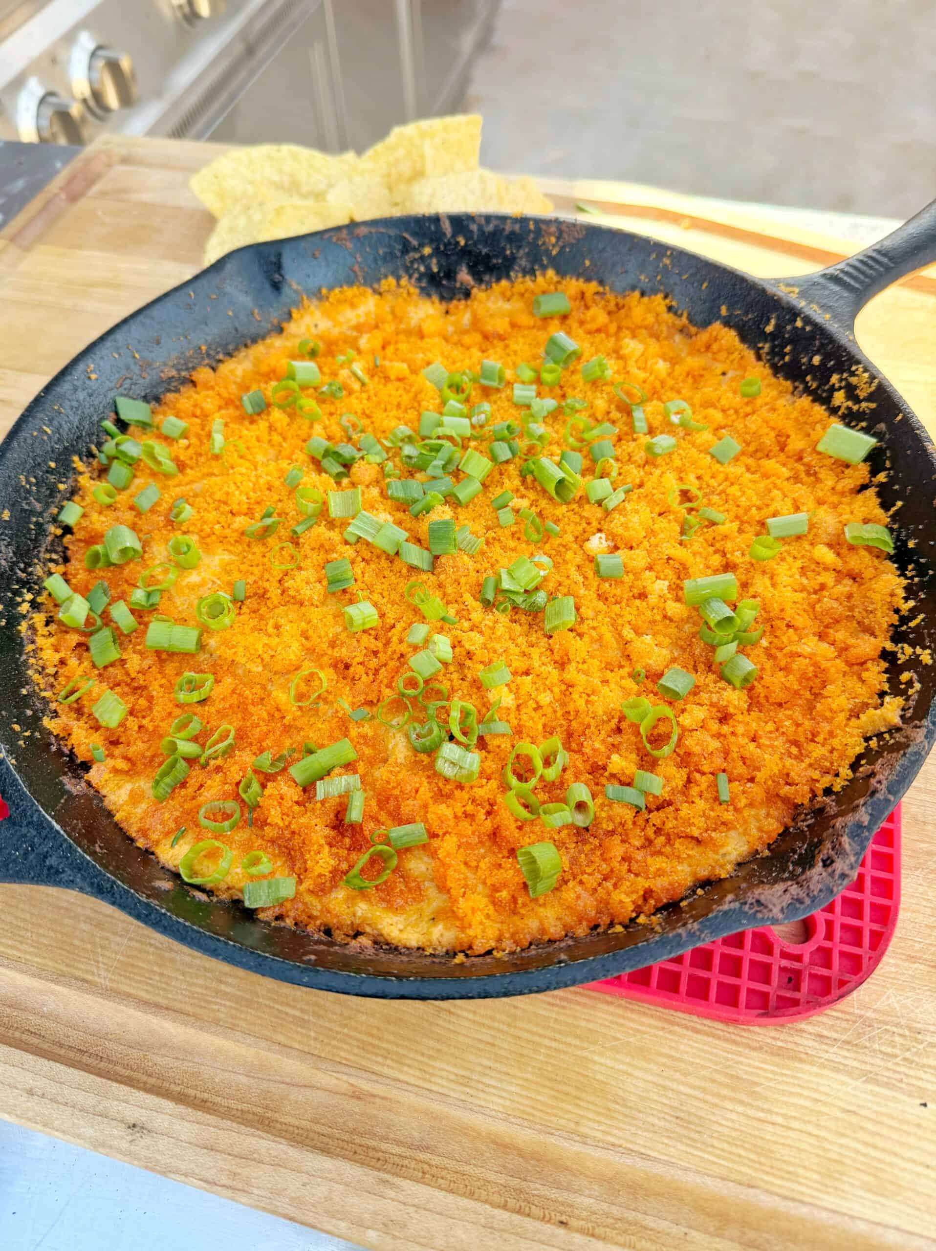 A cast iron skillet filled with a baked dish topped with orange breadcrumbs and chopped green onions, sitting on a wooden surface with tortilla chips and a red hot pad nearby.