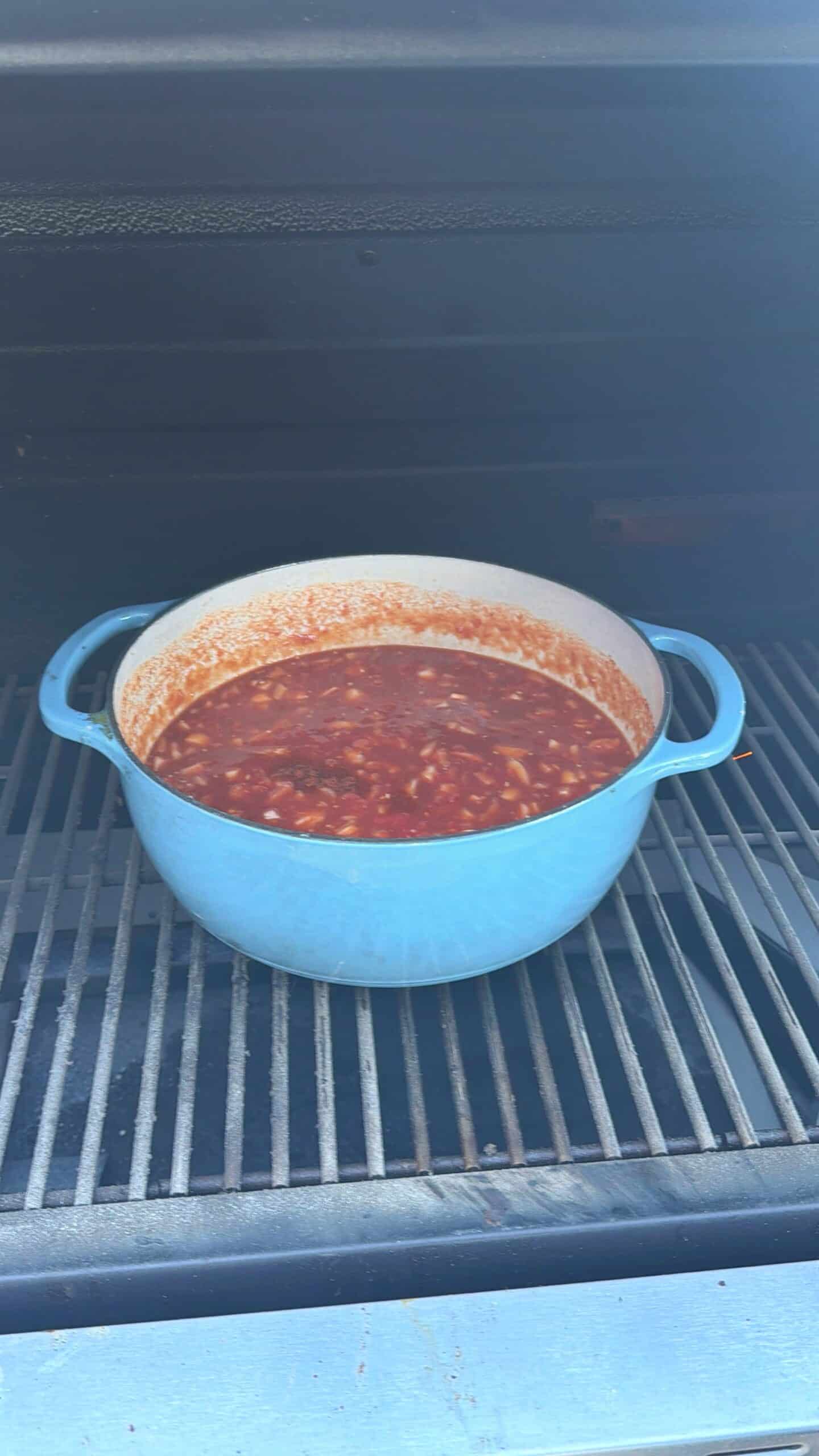 A blue Dutch oven filled with red sauce and shredded meat sits on the grate inside a grill, cooking with the grill lid open.