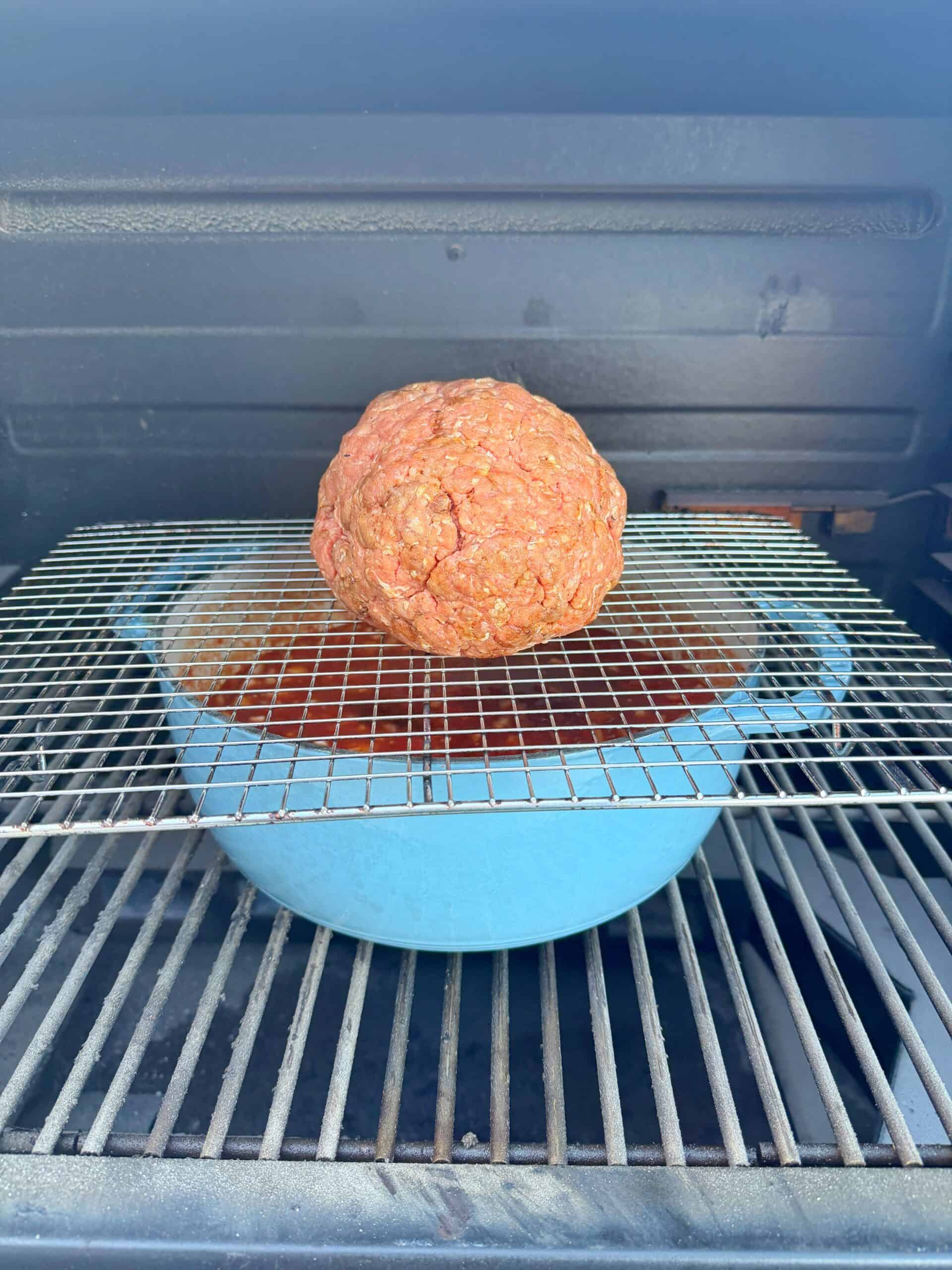A large ball of raw ground meat sits on a wire rack above a blue pot, inside a smoker or grill. The pot appears to catch drippings, and the scene suggests the start of a meat-smoking or roasting process.
