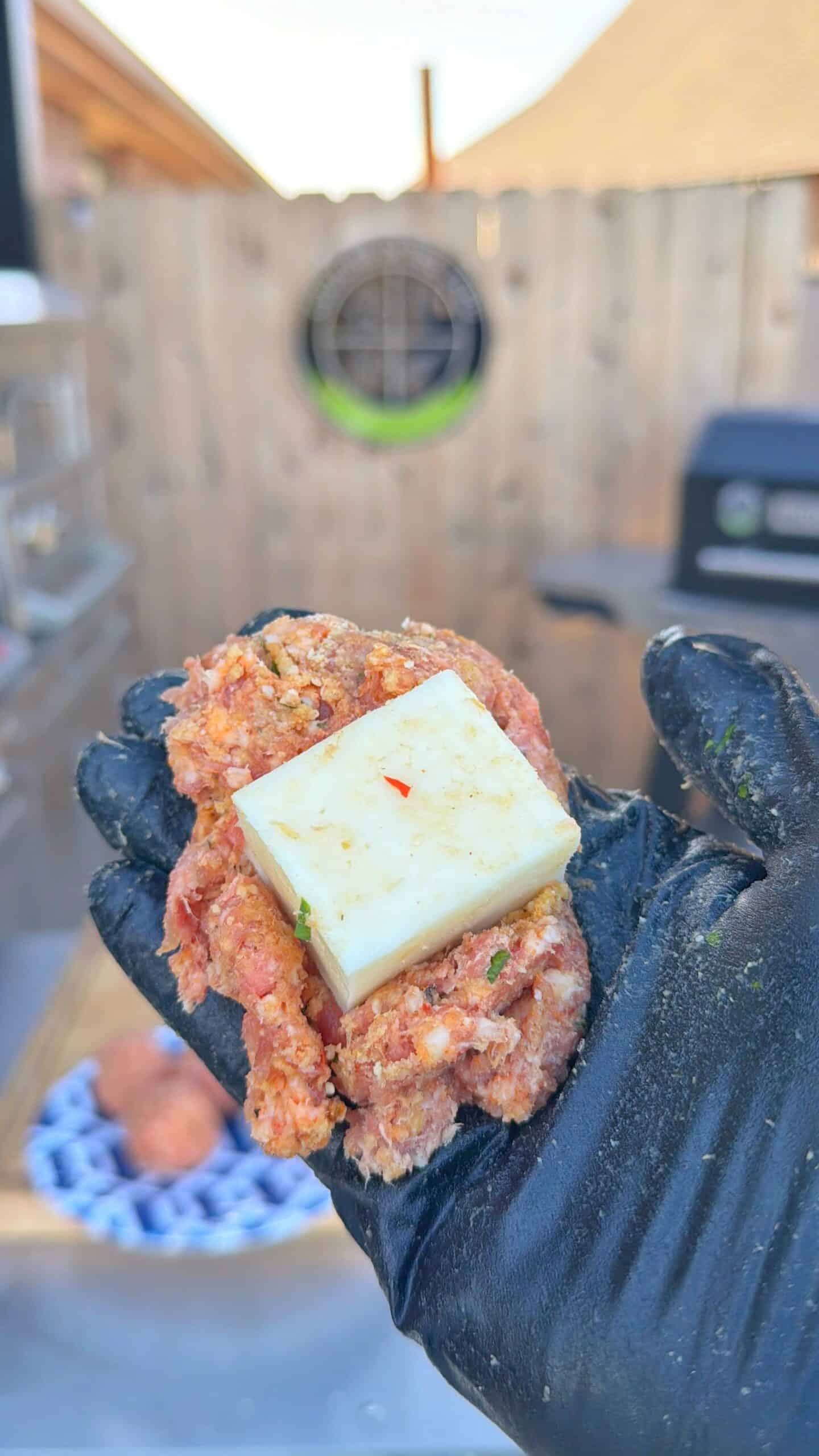 A gloved hand holds seasoned ground meat topped with a thick square slice of cheese, preparing for grilling outdoors near a wooden fence and barbecue grill making a stuffed meatball.
