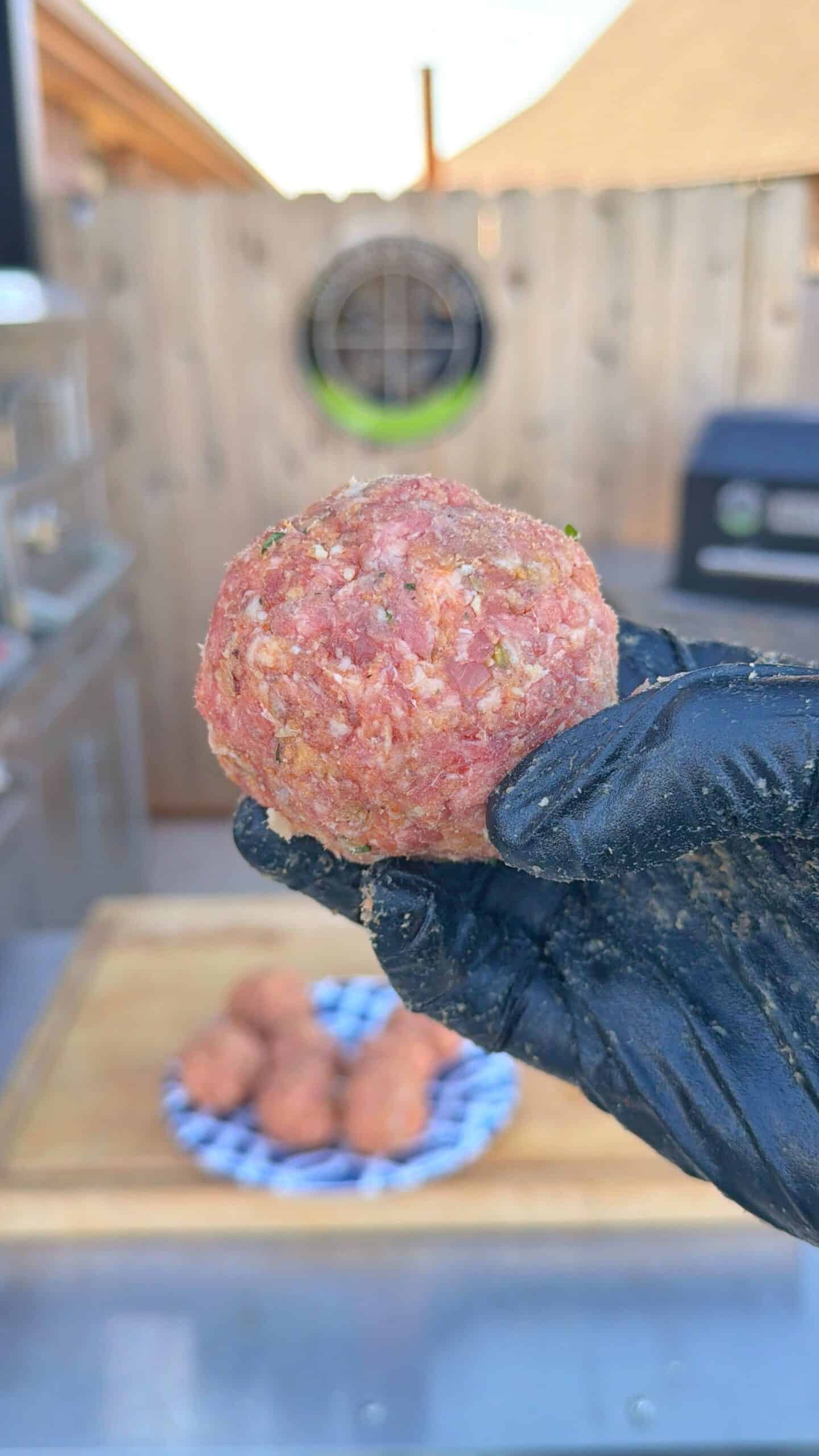 A gloved hand holds a seasoned raw meatball with more meatballs on a plate in the background, set in an outdoor kitchen area with a grill and wooden fence visible.