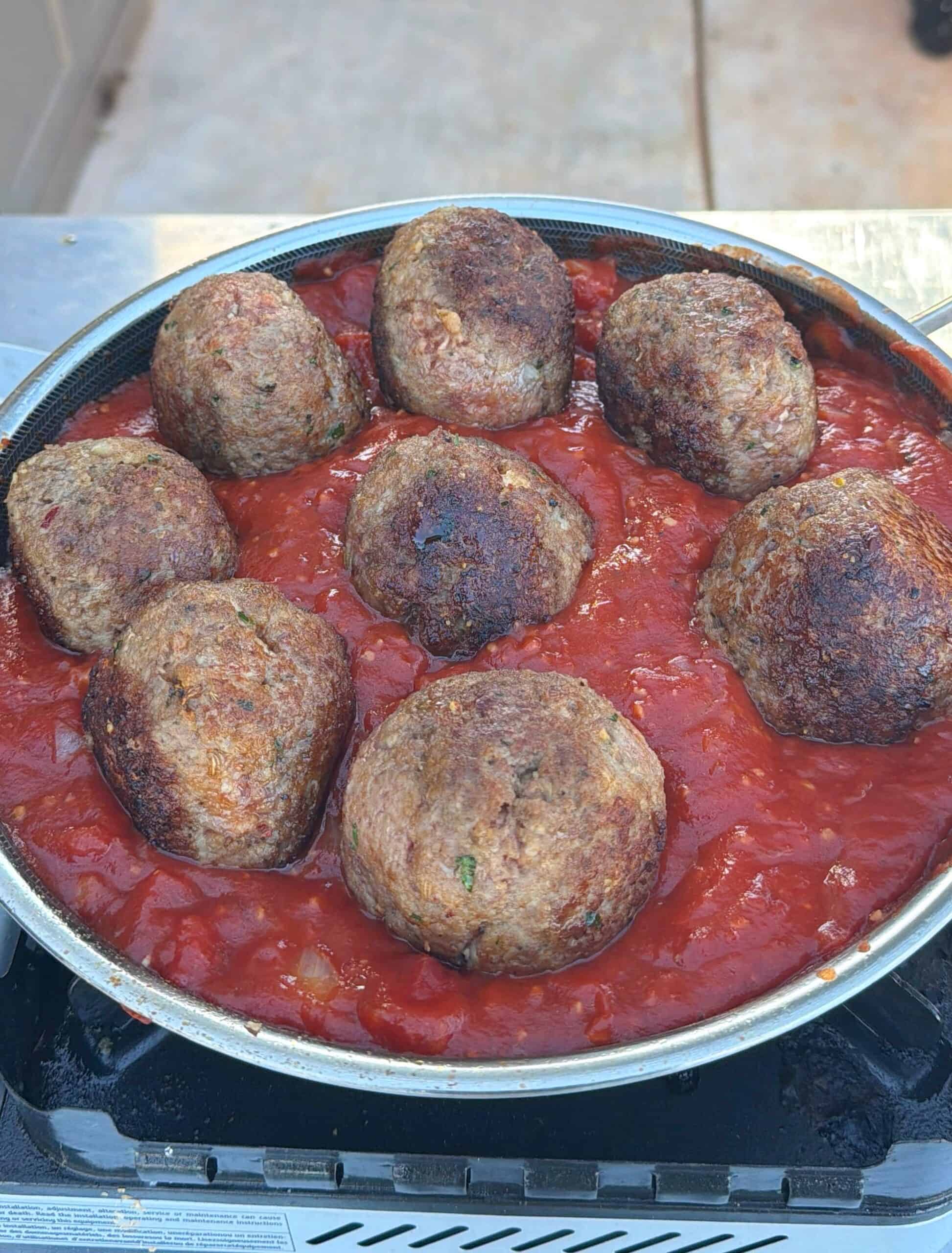 A pan filled with chunky tomato sauce and eight large, browned meatballs cooking on top, set on an outdoor stove.