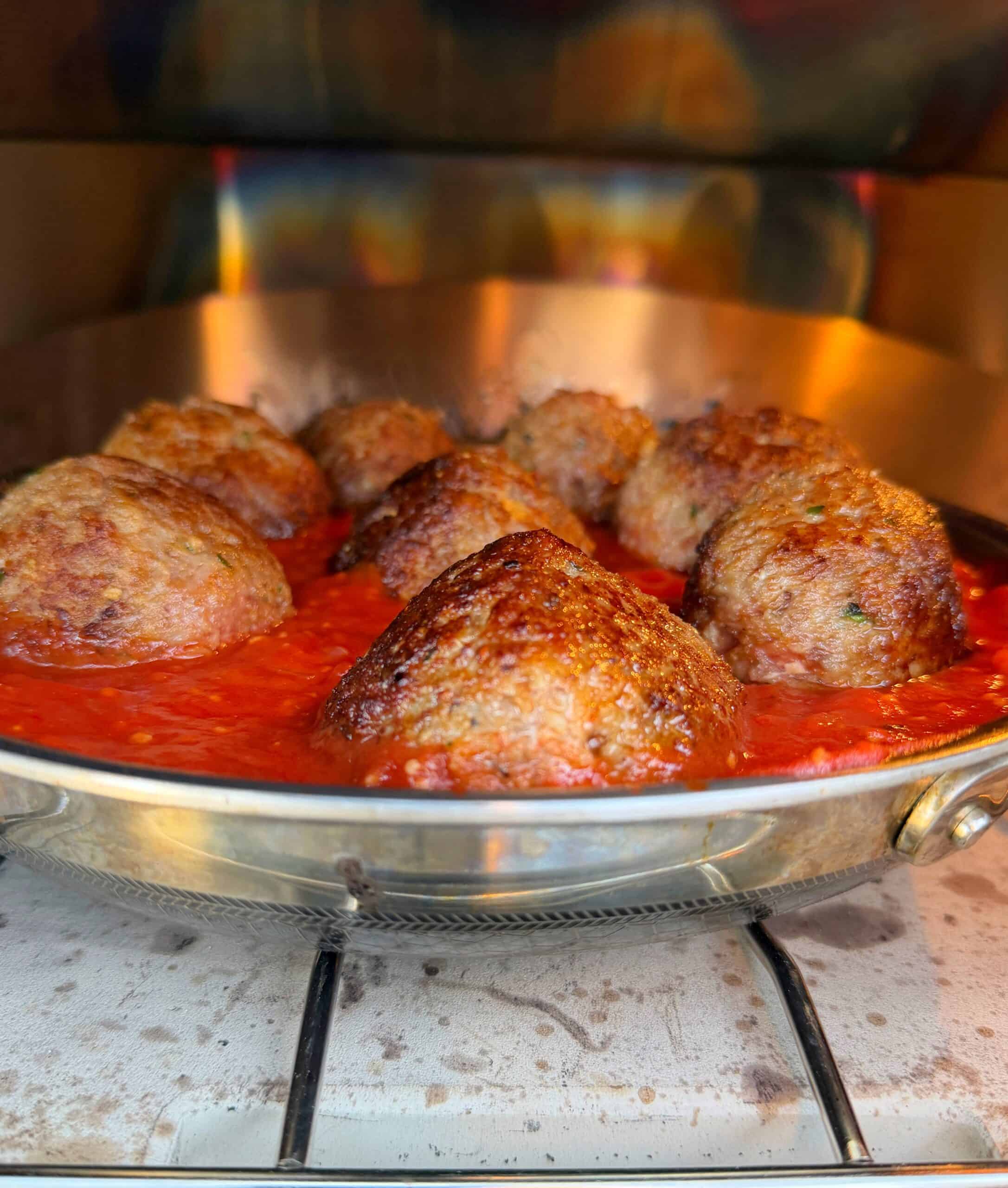 A close-up of several browned meatballs simmering in red tomato sauce in a metal pan on a stovetop.