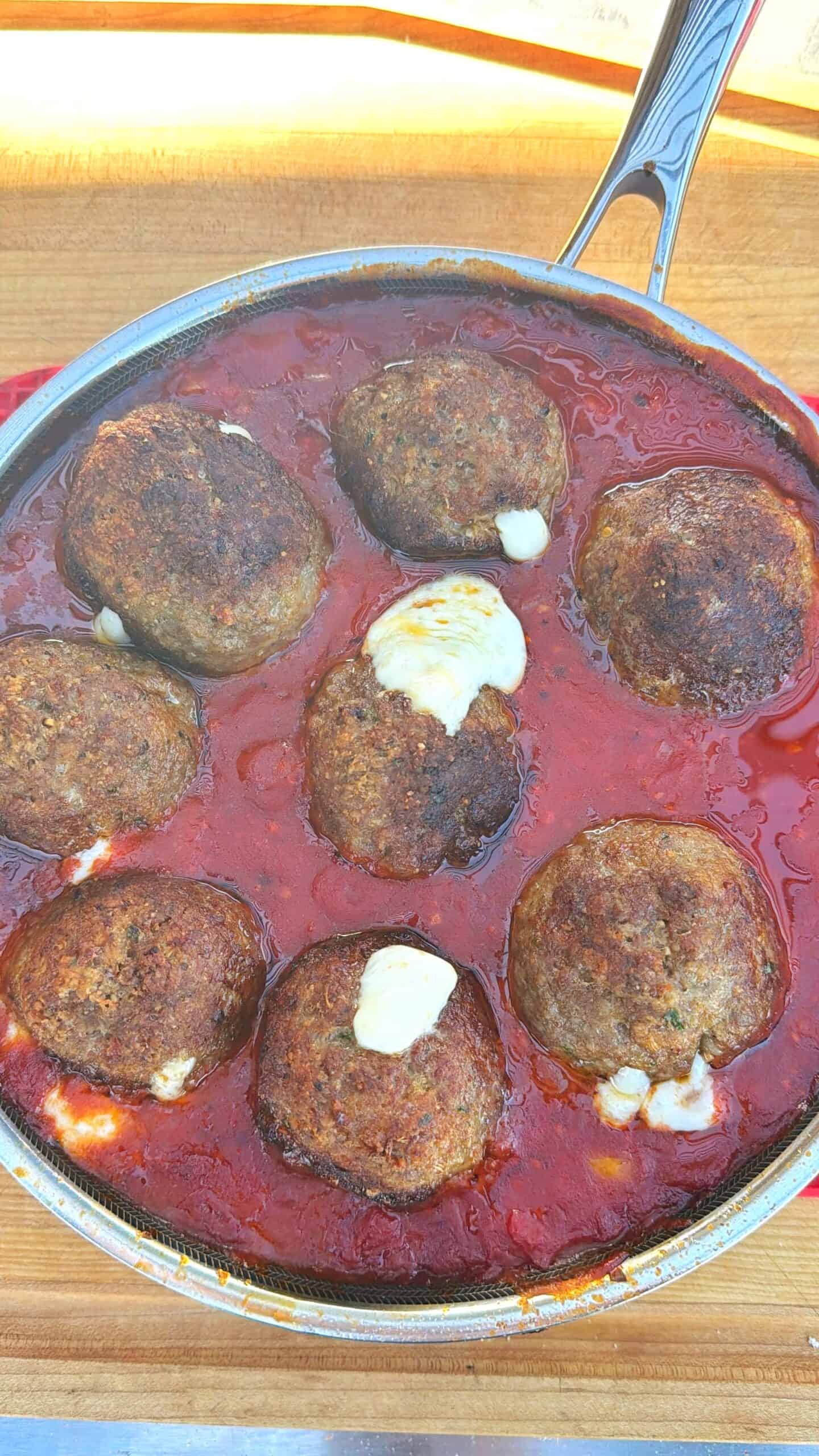 A skillet filled with large meatballs partially covered in melted cheese, simmering in rich red tomato sauce on a wooden surface.