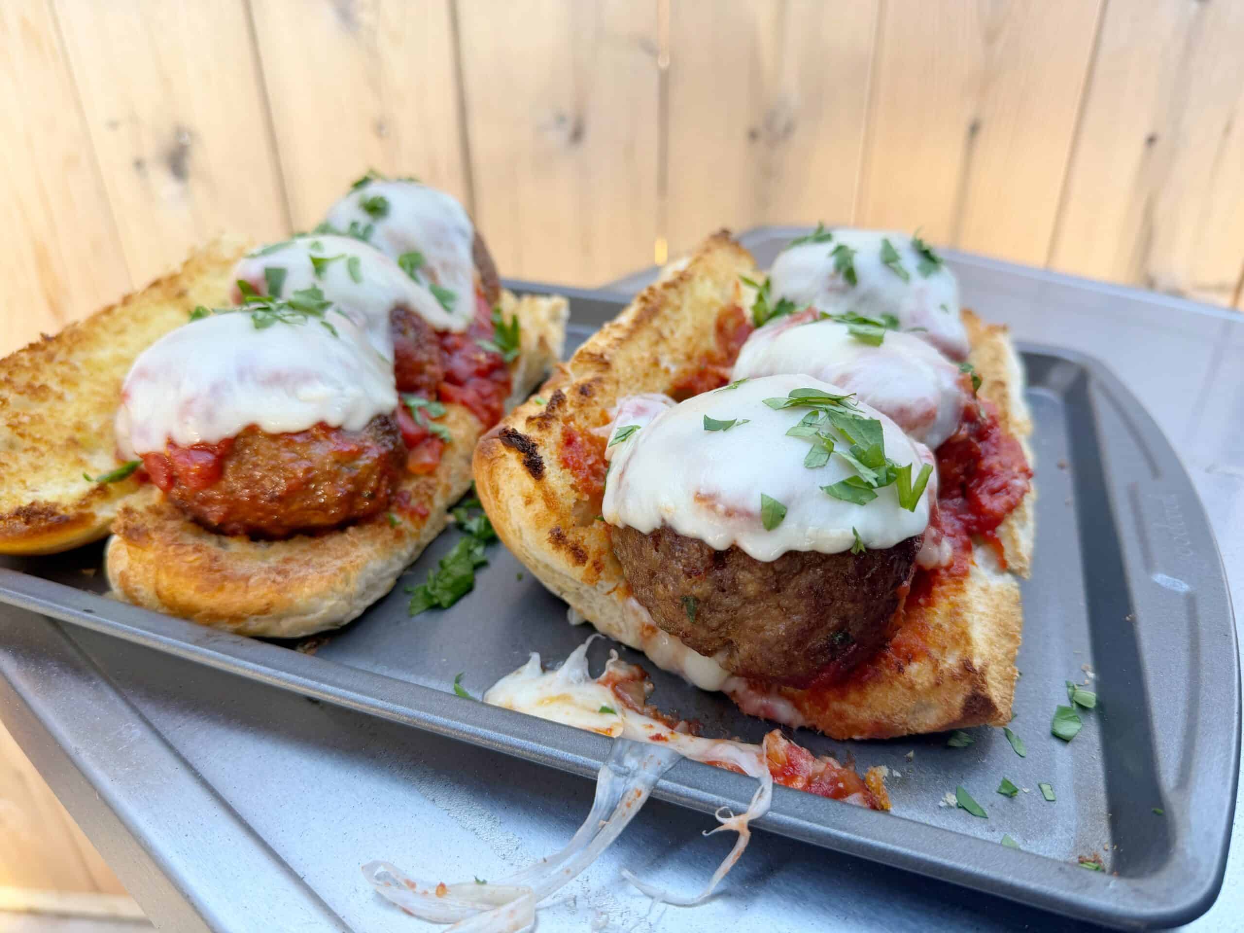 Two toasted hoagie rolls filled with large meatballs, marinara sauce, and melted cheese, garnished with chopped herbs, sit on a metal baking tray with cheese and sauce spilling onto the tray.