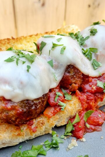 A toasted sub roll topped with meatballs, marinara sauce, melted mozzarella cheese, and fresh chopped parsley, served on a metal tray with a wooden background.