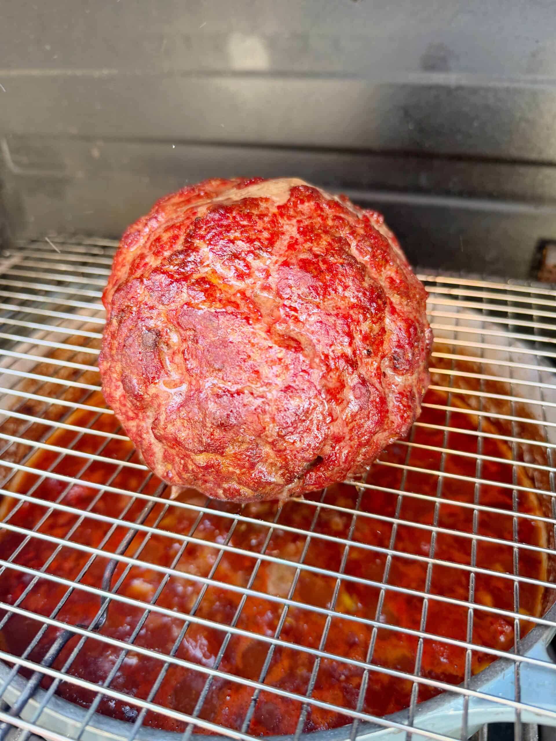 A round, cooked meatloaf with a reddish glaze sits on a metal cooling rack above a pan catching juices inside a grill or smoker.