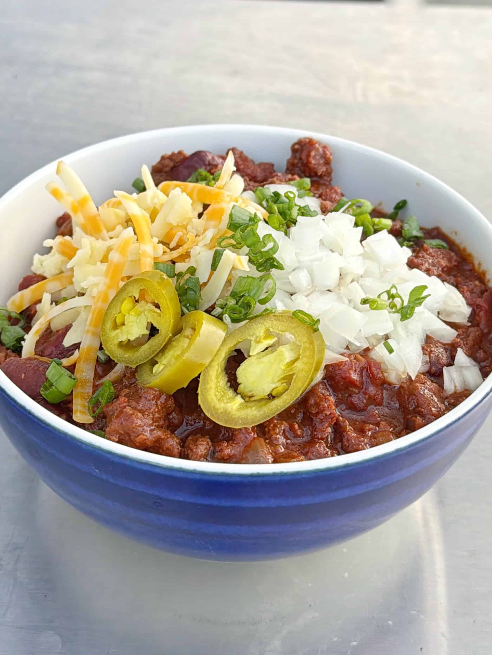 A bowl of chili topped with shredded cheese, chopped onions, sliced jalapeños, and green onions, served in a blue and white bowl on a light surface.