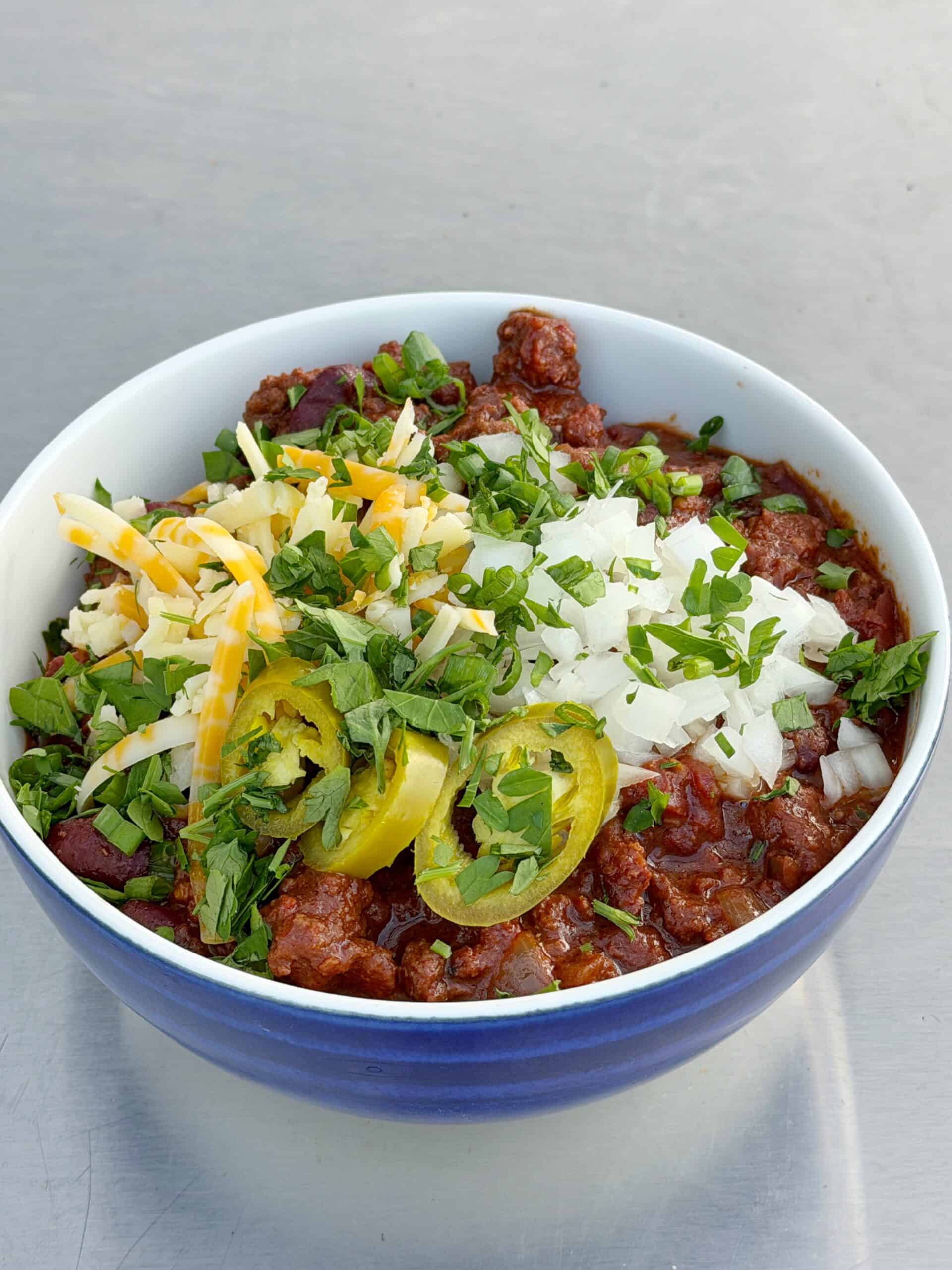 A bowl of chili topped with shredded cheese, chopped onions, sliced jalapeños, and fresh herbs, set on a light gray surface.