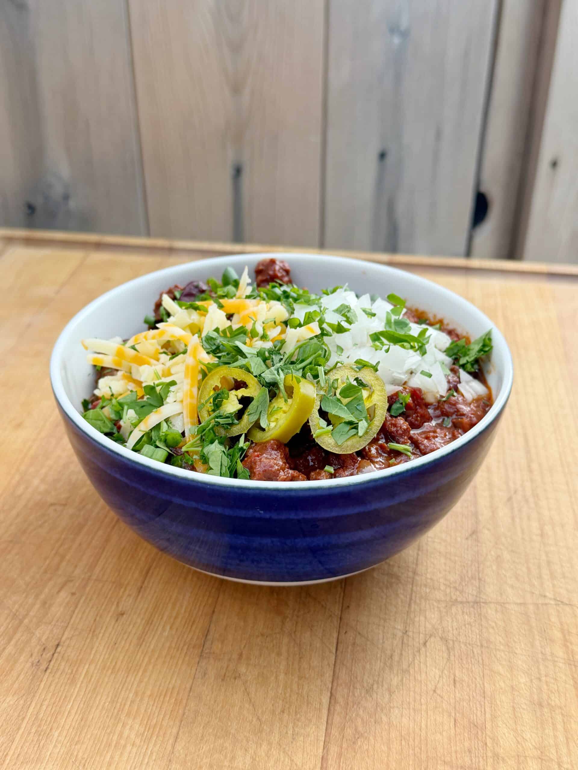 A bowl of chili topped with shredded cheese, chopped onions, sliced jalapeños, and fresh herbs sits on a wooden surface with a light wood background.