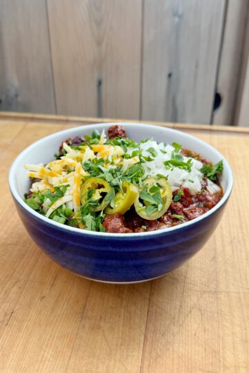 A bowl of chili topped with shredded cheese, chopped onions, sliced jalapeños, and fresh herbs, sitting on a wooden surface with a wood-paneled background.
