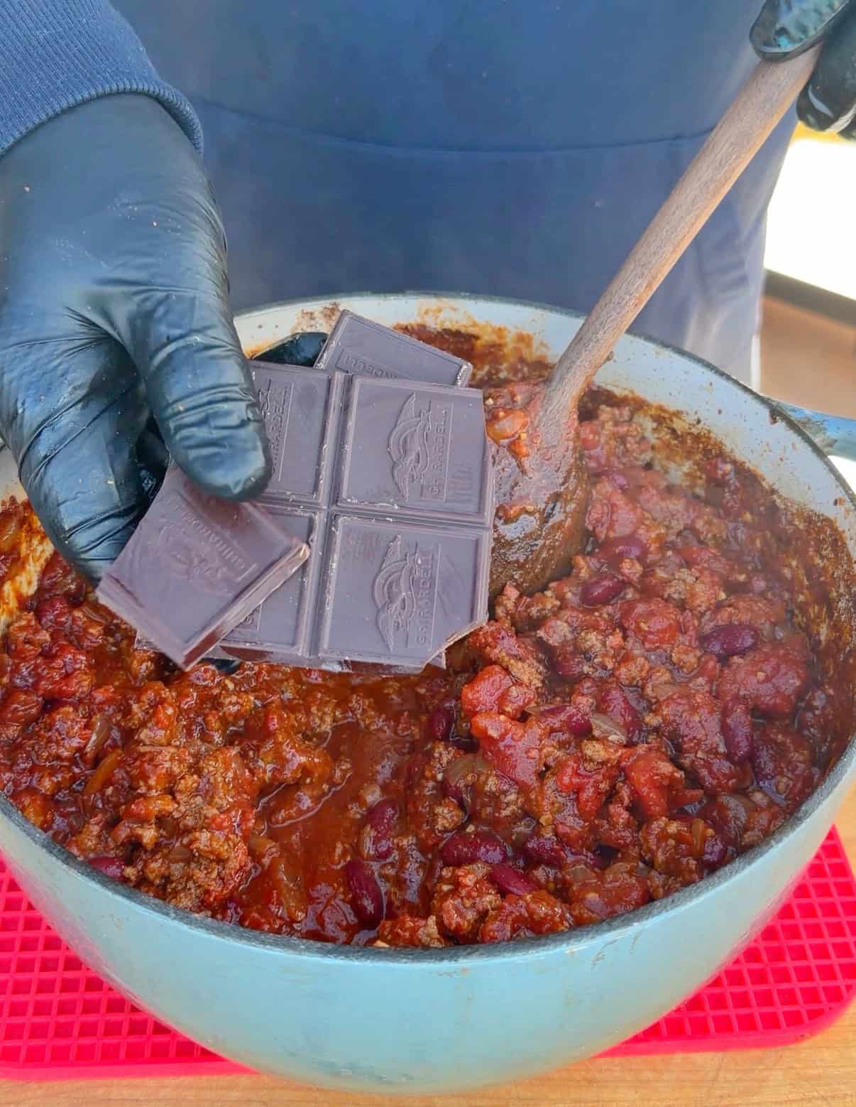 A hand in a black glove adds a bar of dark chocolate to a pot of chili with beans and ground meat, using a wooden spoon to stir. The pot sits on a red trivet.