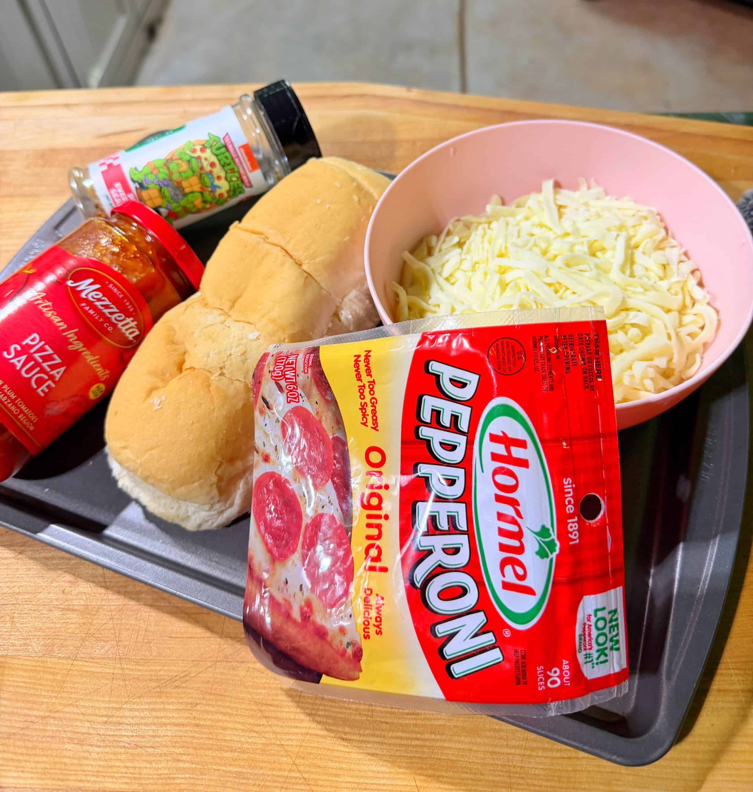 A tray with sub rolls, a bowl of shredded mozzarella, a package of Hormel pepperoni, a jar of pizza sauce, and a container of Italian seasoning on a wooden counter.