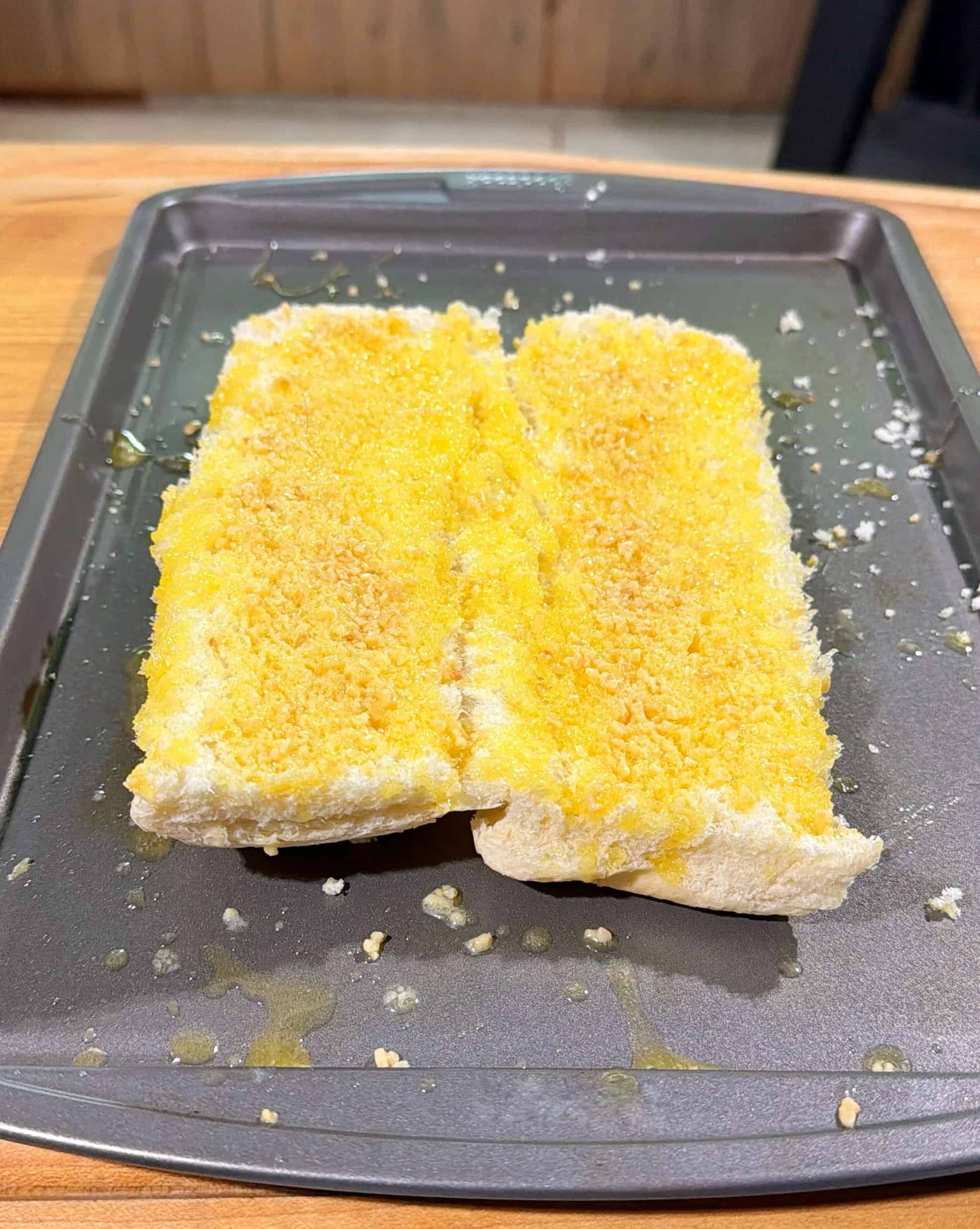 Two slices of bread on a baking tray, spread with yellow melted butter and sprinkled with breadcrumbs, ready to be baked. The tray sits on a wooden surface.