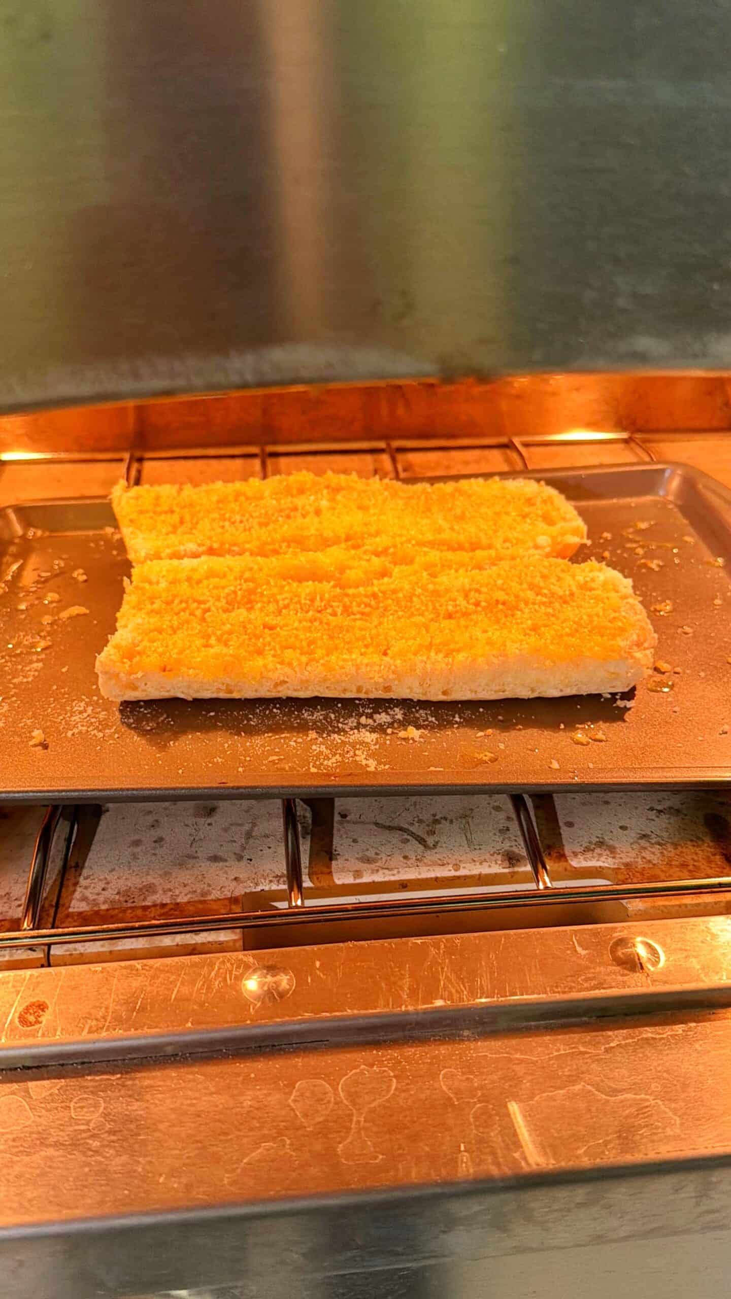 Two breaded fish fillets are baking on a metal tray inside an oven. The fillets are golden and crispy, sitting side by side on the tray above the oven rack.