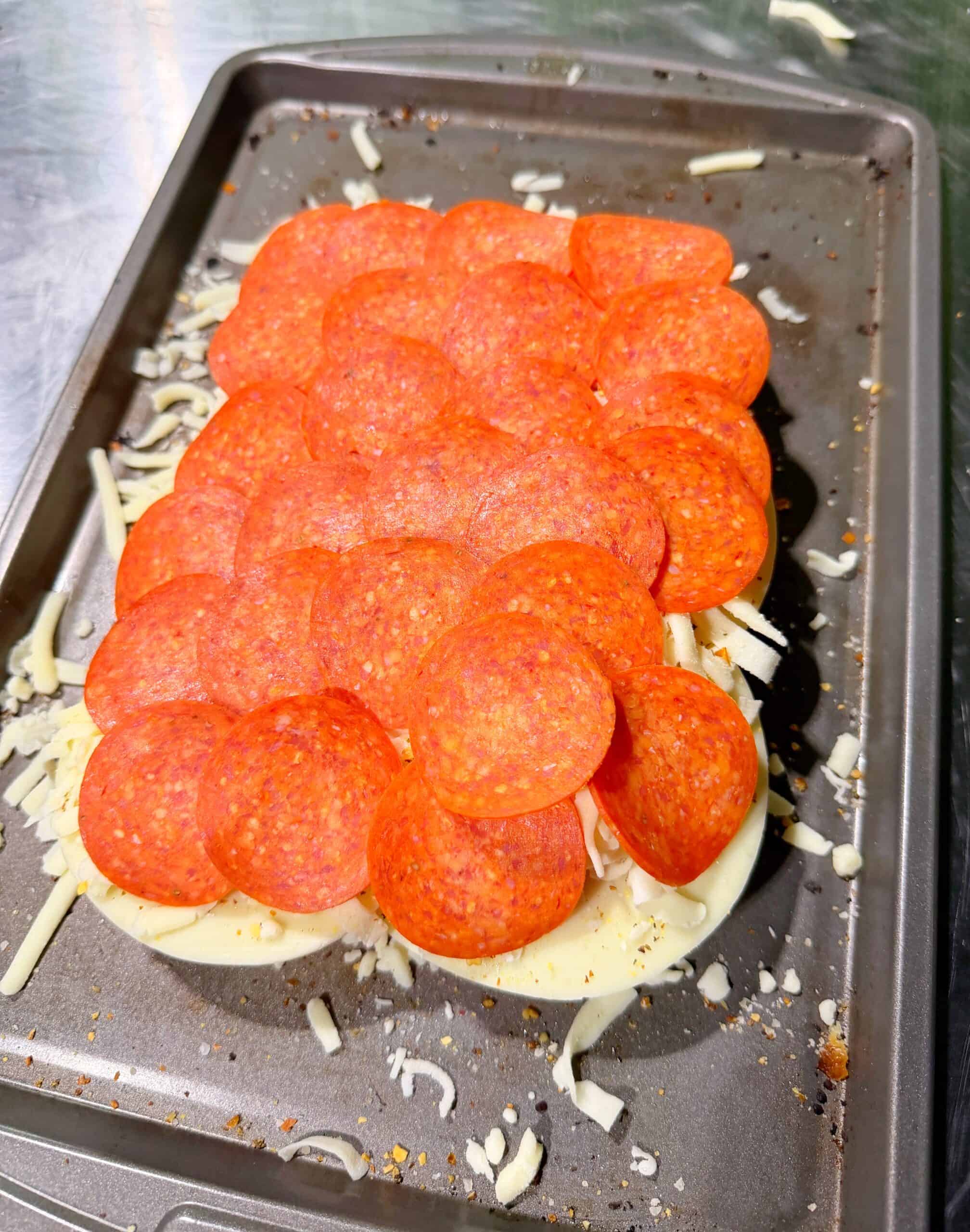 A baking tray holding an uncooked dish topped with layers of shredded cheese and overlapping slices of pepperoni, ready to be baked. Some cheese is scattered around the tray.