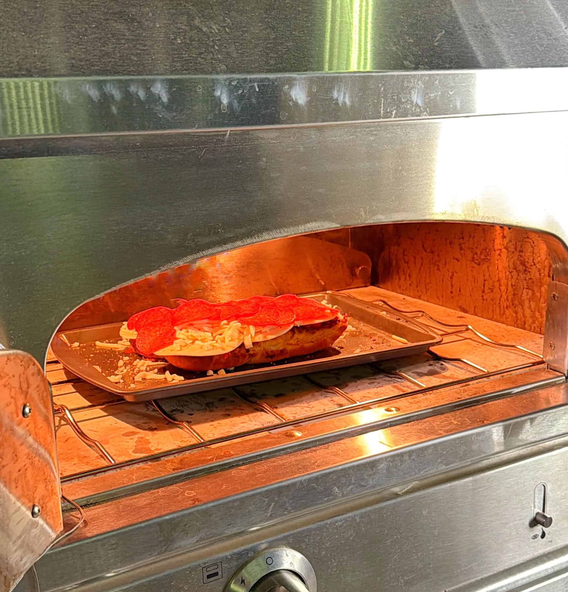 A pizza topped with pepperoni and cheese is baking on a metal tray inside a stainless steel pizza oven. Warm light from the oven illuminates the pizza and the oven’s interior.