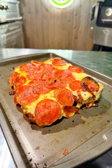 A rectangular pepperoni pizza with melted cheese sits on a metal baking sheet. The background shows a stainless steel kitchen and wood-paneled wall. The pizza edges are crispy and golden brown.
