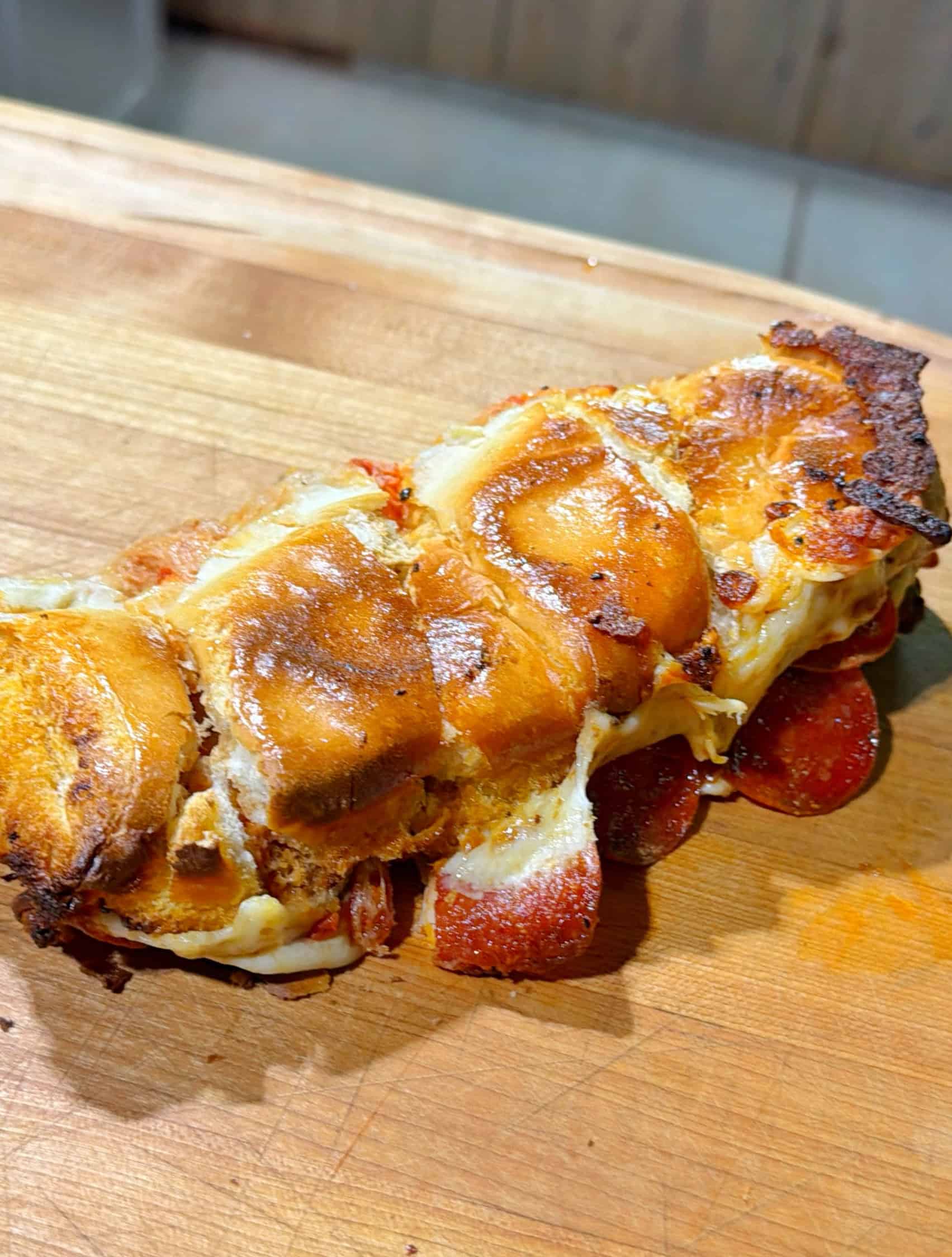 A close-up of a baked sandwich on a wooden cutting board, filled with melted cheese and pepperoni, with a golden-brown, crispy crust.