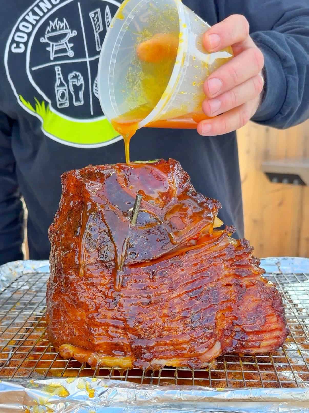 A person pours a golden glaze over a sliced, smoked ham resting on a wire rack covered with foil. The person wears a black shirt with a grilling-themed logo. The scene appears to be outdoors.