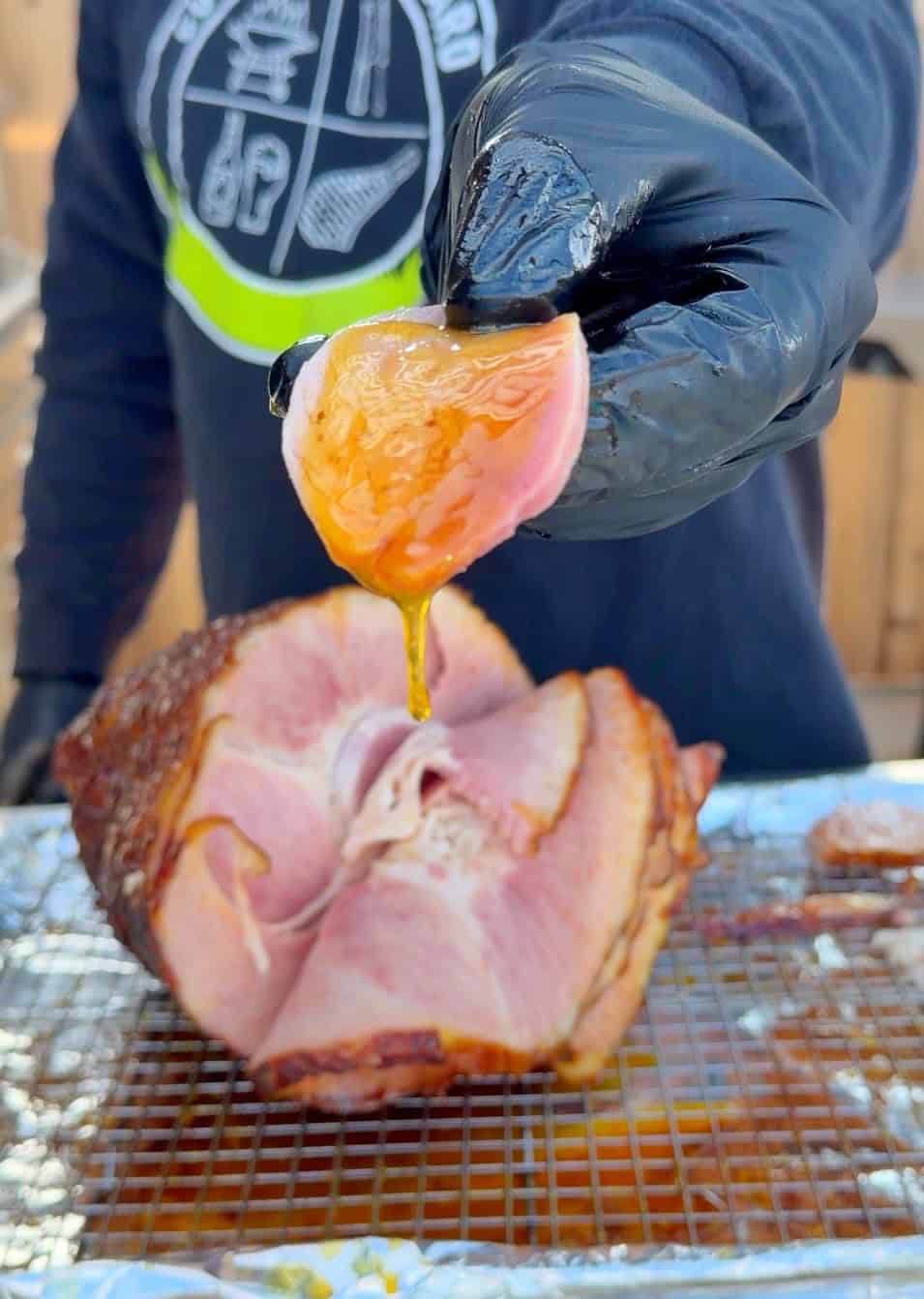 A gloved hand holds a slice of glazed ham with honey dripping from it, in front of a whole ham resting on a foil-covered rack. The person wears a dark sweatshirt featuring a bold graphic design.