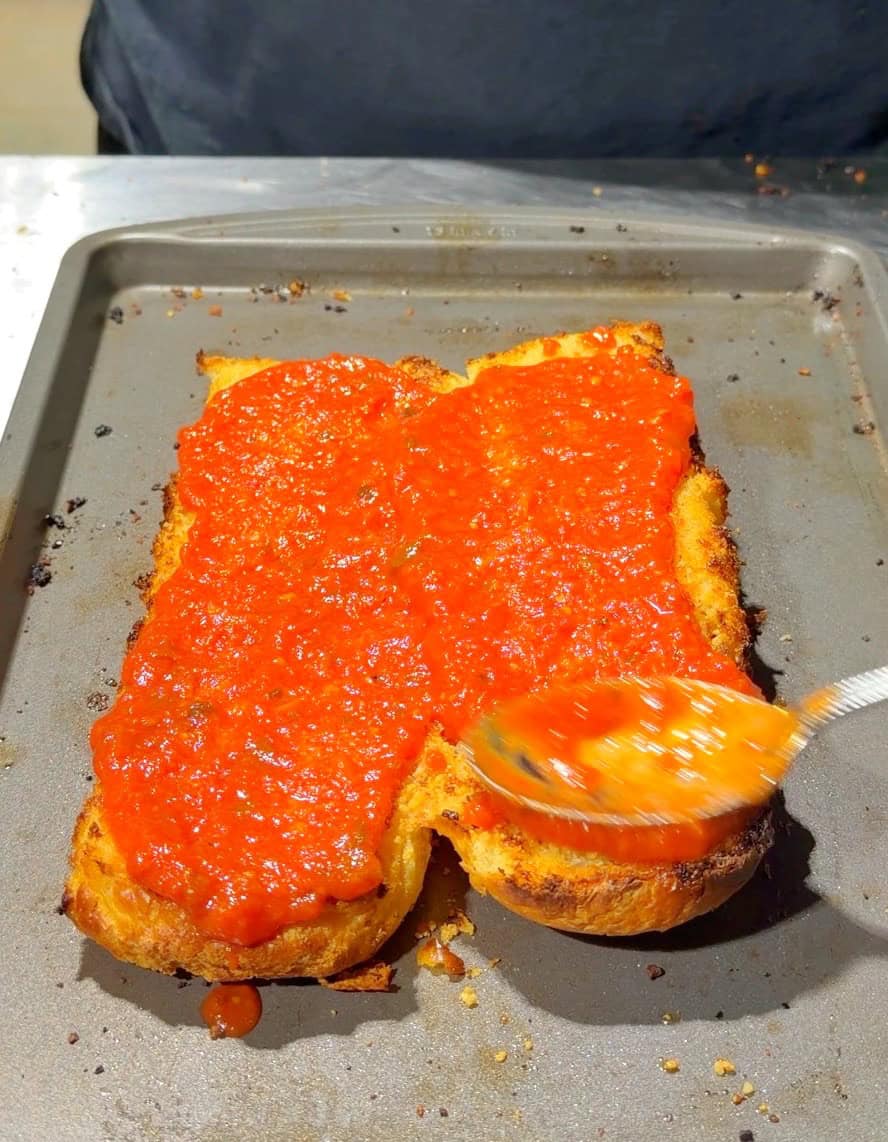 A close-up of a sheet pan with toasted bread topped with red tomato sauce. A hand is spreading the sauce with a spoon, and the background is out of focus.