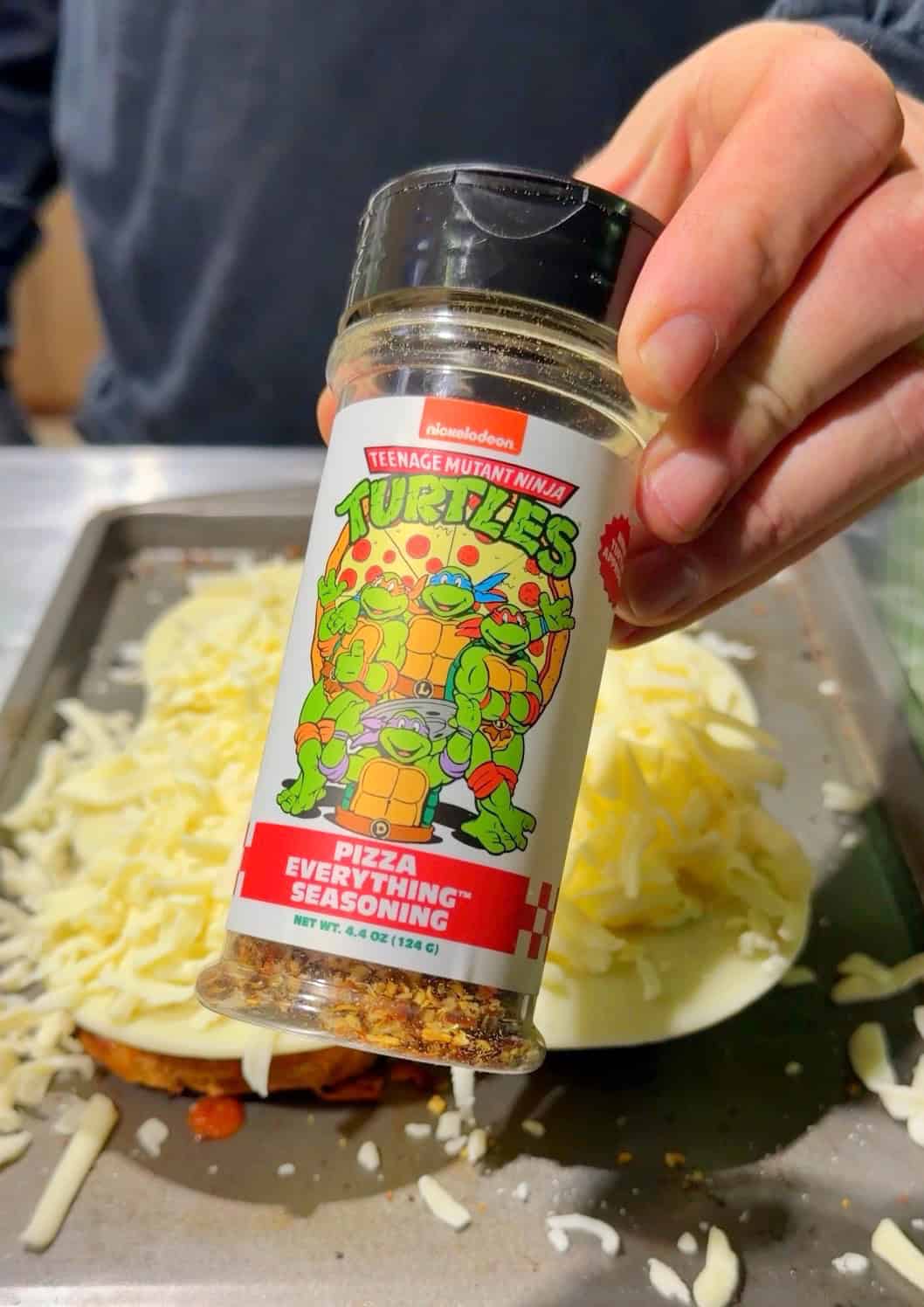 A hand holds a Teenage Mutant Ninja Turtles Pizza Everything seasoning bottle over a tray with cheese-covered food, ready to be baked.