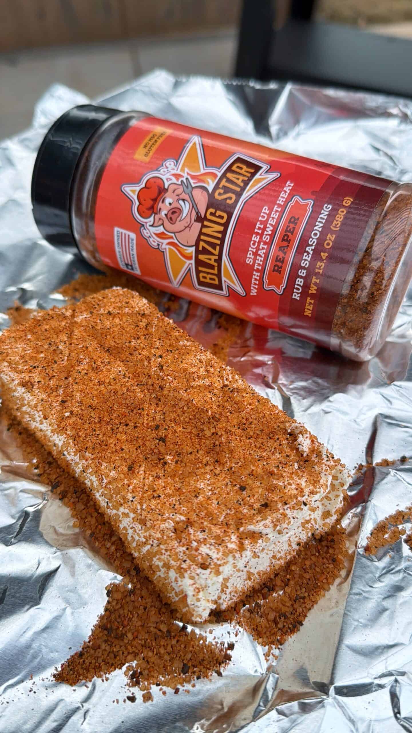 A block of white cheese is covered in a reddish-brown seasoning, lying on aluminum foil. Next to it is a bottle of Blazing Star Reaper hot seasoning, with some spilled on the foil.