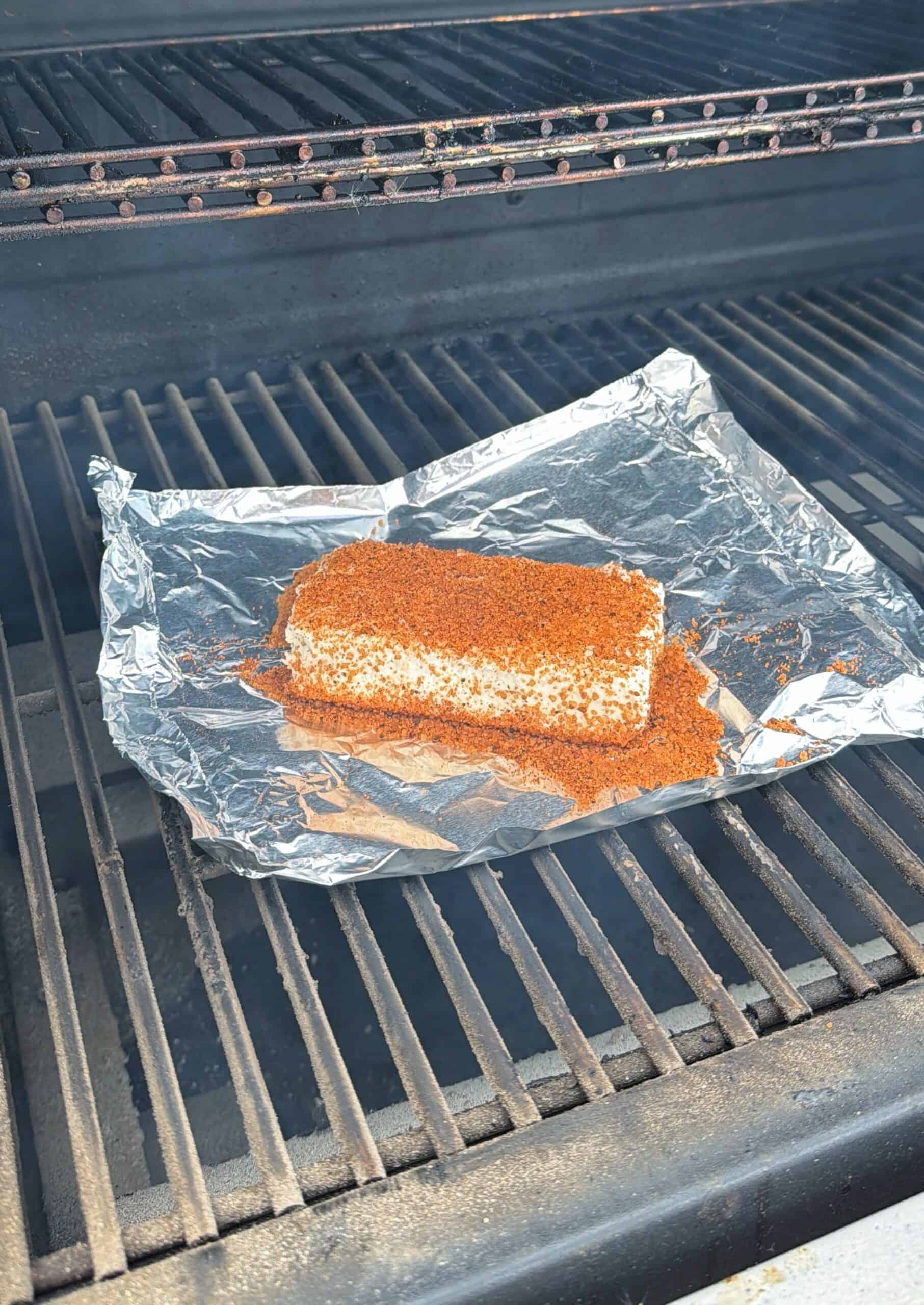 A block of cream cheese coated with seasoning sits on a piece of aluminum foil placed on the grates of a grill. The grill lid is open, and the cream cheese is ready to be smoked or cooked.