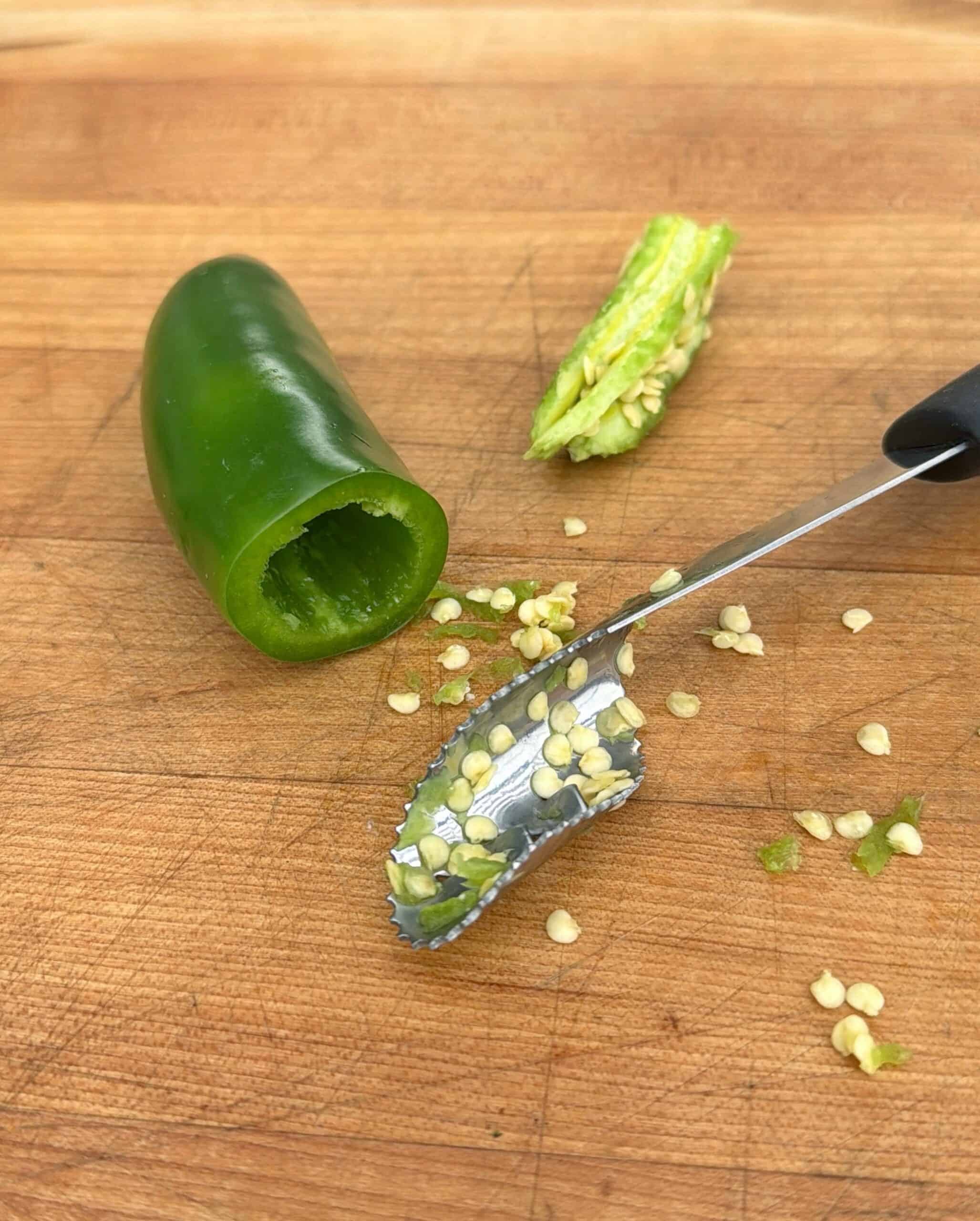 A partially hollowed jalapeño pepper, its top and seeds removed, sits on a wooden cutting board. A small metal tool with seeds on it rests nearby.