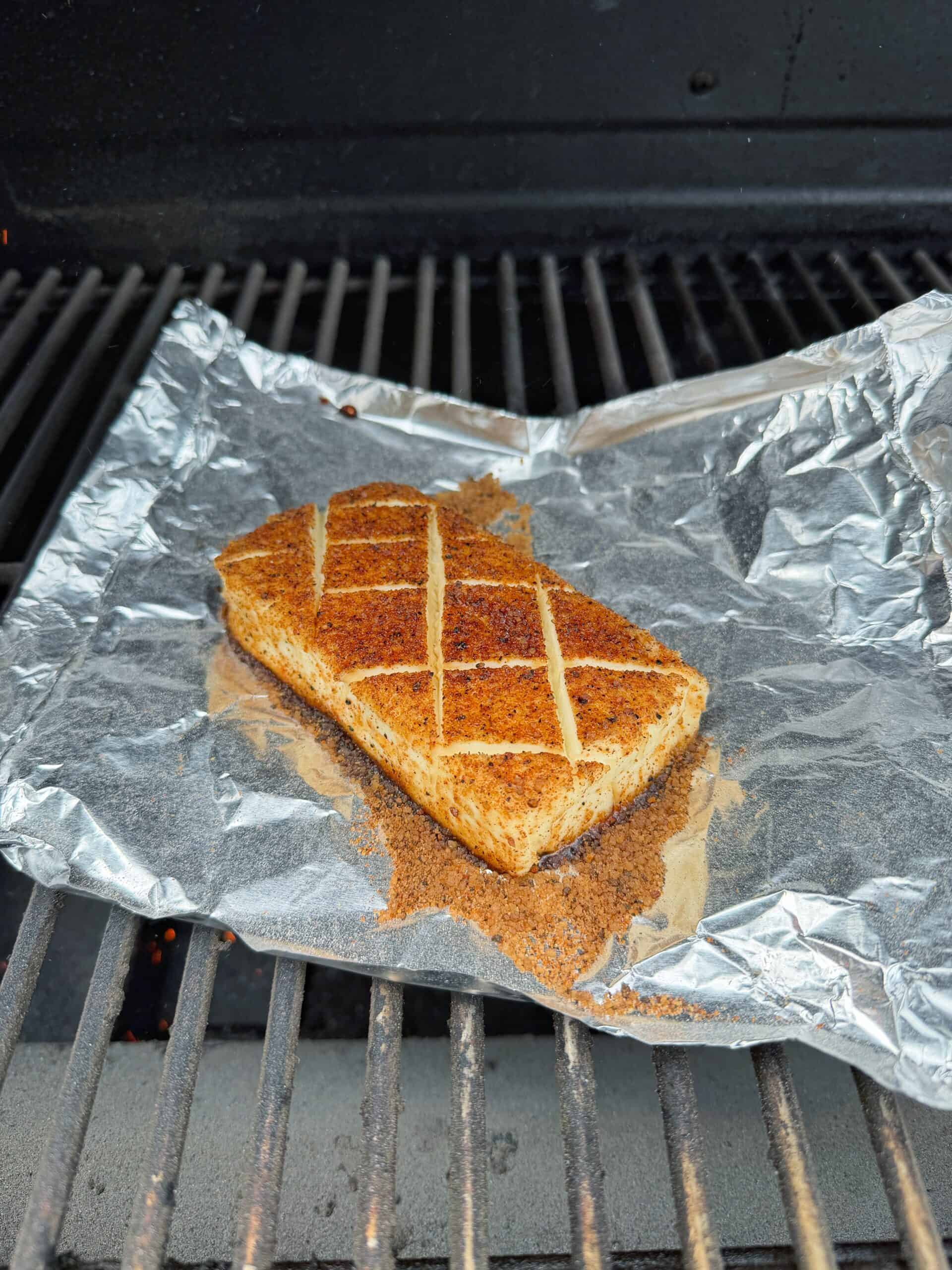A seasoned block of tofu with crisscross knife marks is grilling on a piece of foil on a barbecue grill.