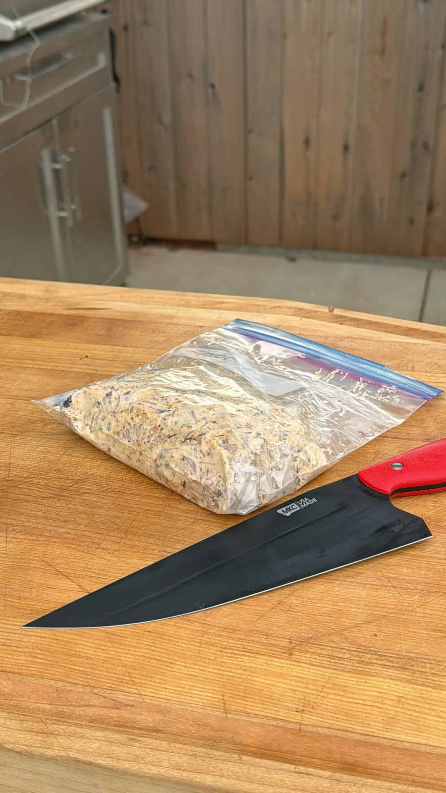 A large black chef’s knife with a red handle lies on a wooden countertop next to a resealable plastic bag filled with shredded cooked meat. A stainless steel appliance and wooden wall are in the background.