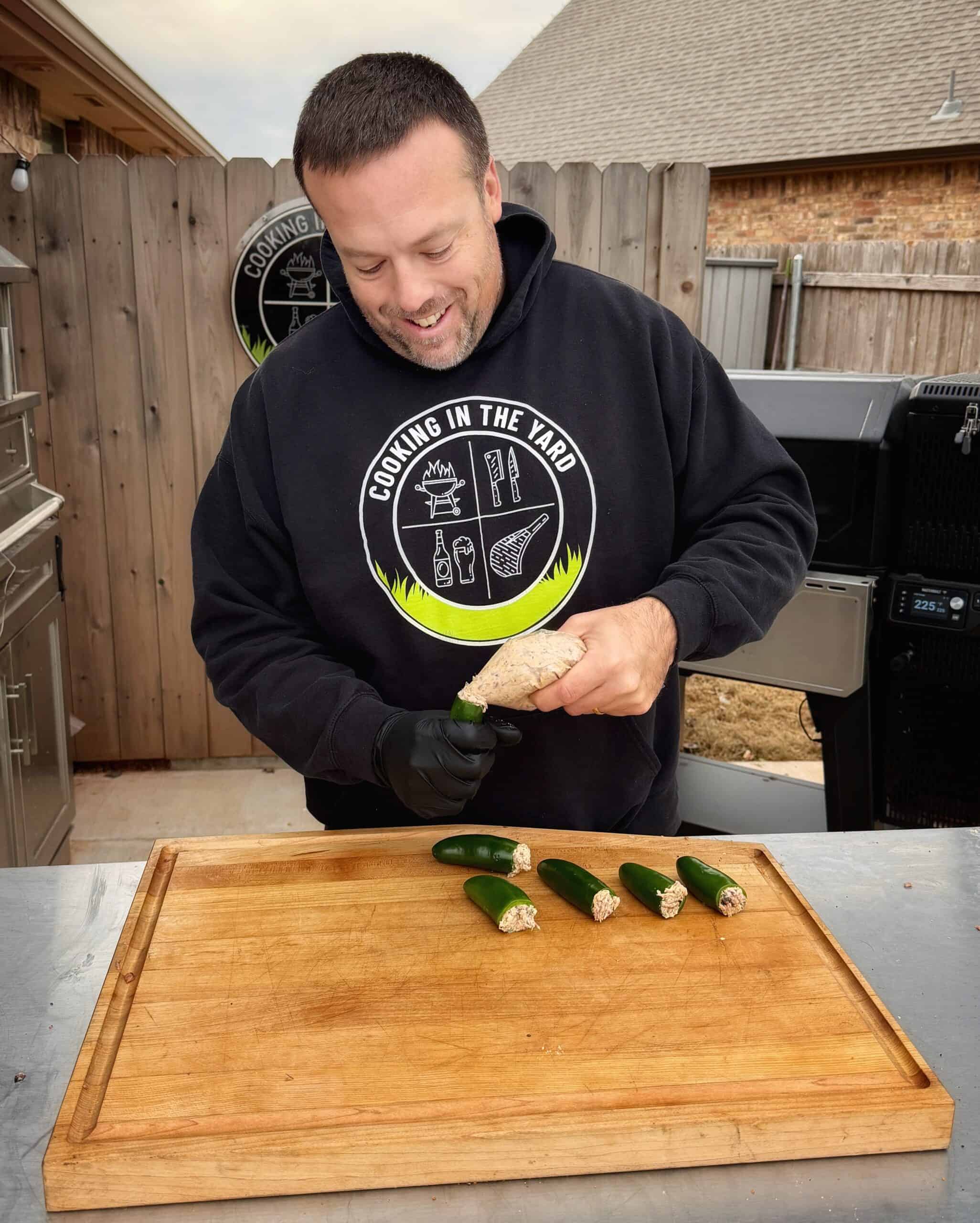 A man wearing a Cooking in the Yard hoodie and black gloves stuffs jalapeños with a filling at an outdoor kitchen, standing in front of a wooden cutting board.