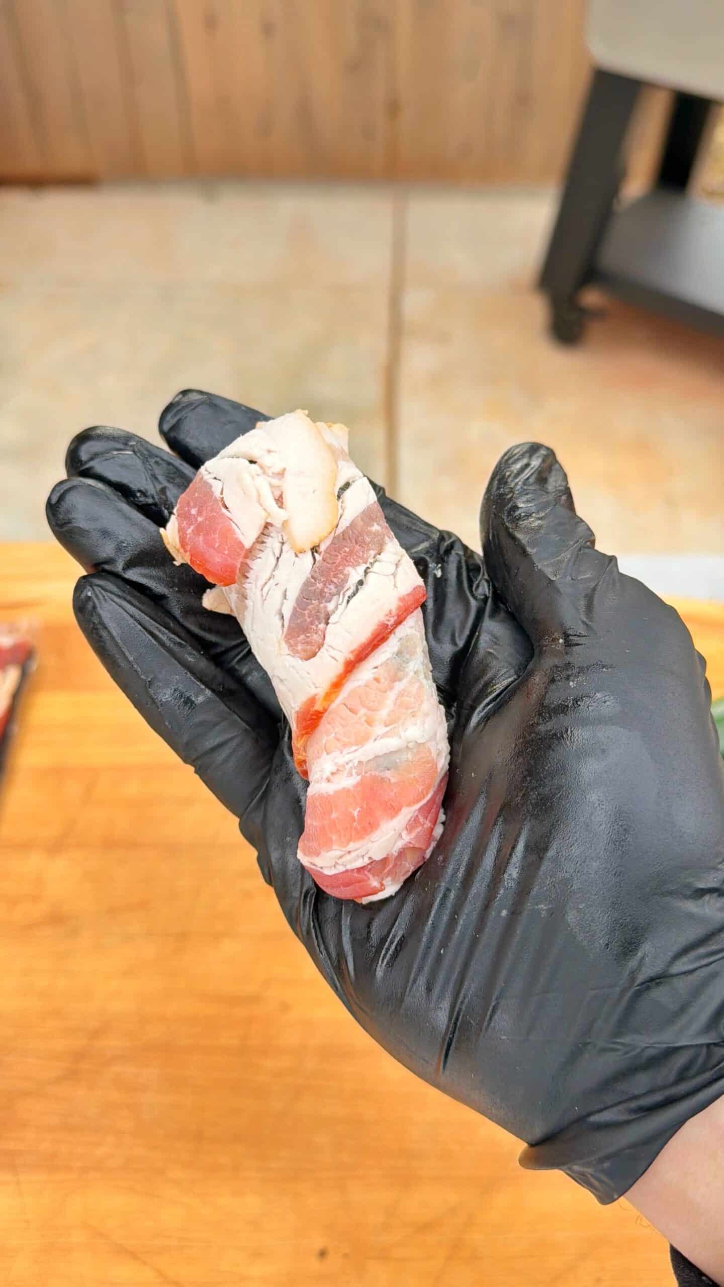 A hand wearing a black glove holds a small piece of raw meat wrapped in bacon, with a wooden surface and grill visible in the background.