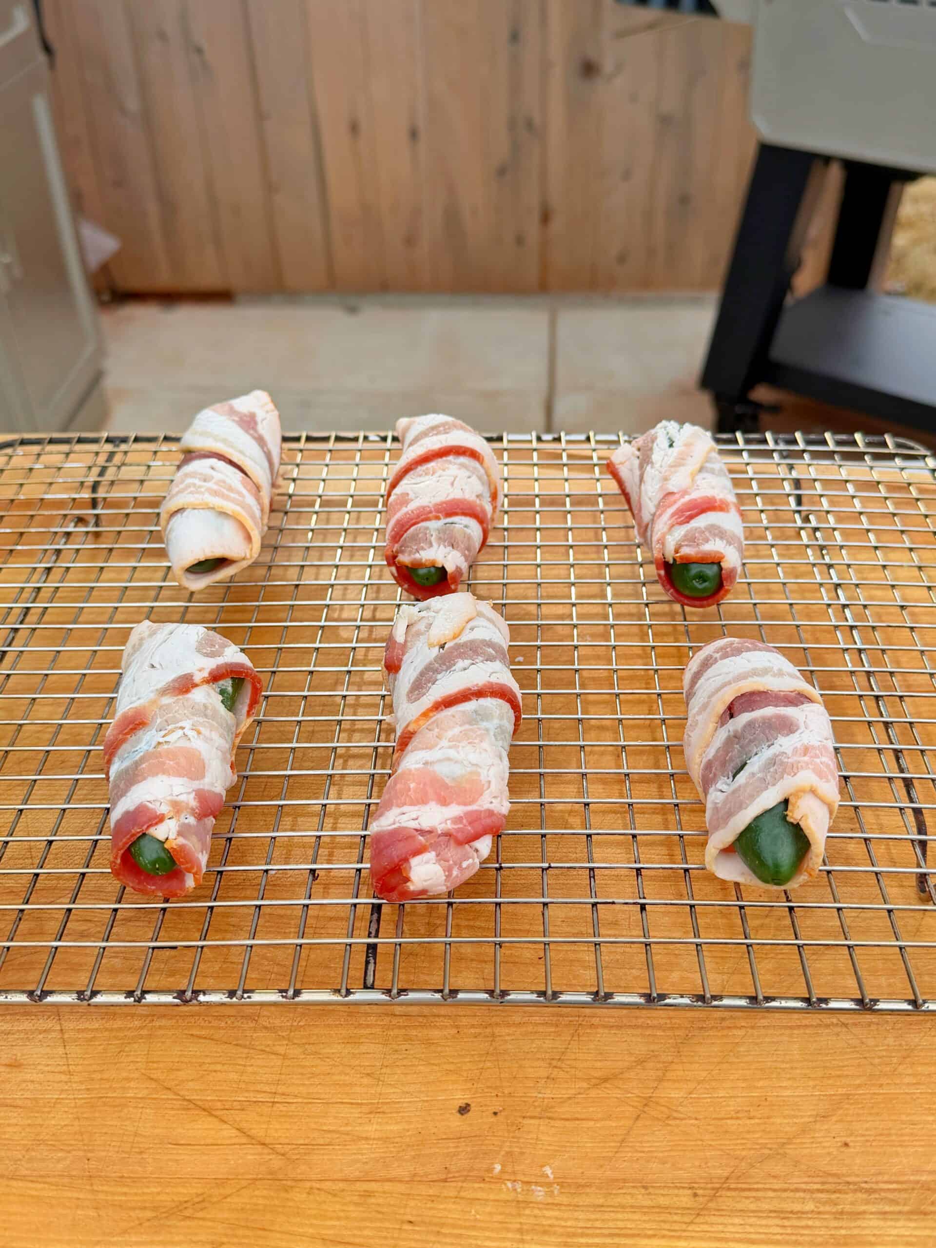Six bacon-wrapped jalapeño poppers arranged on a metal cooling rack over a wooden surface, ready to be cooked. A grill and wooden wall are visible in the background.