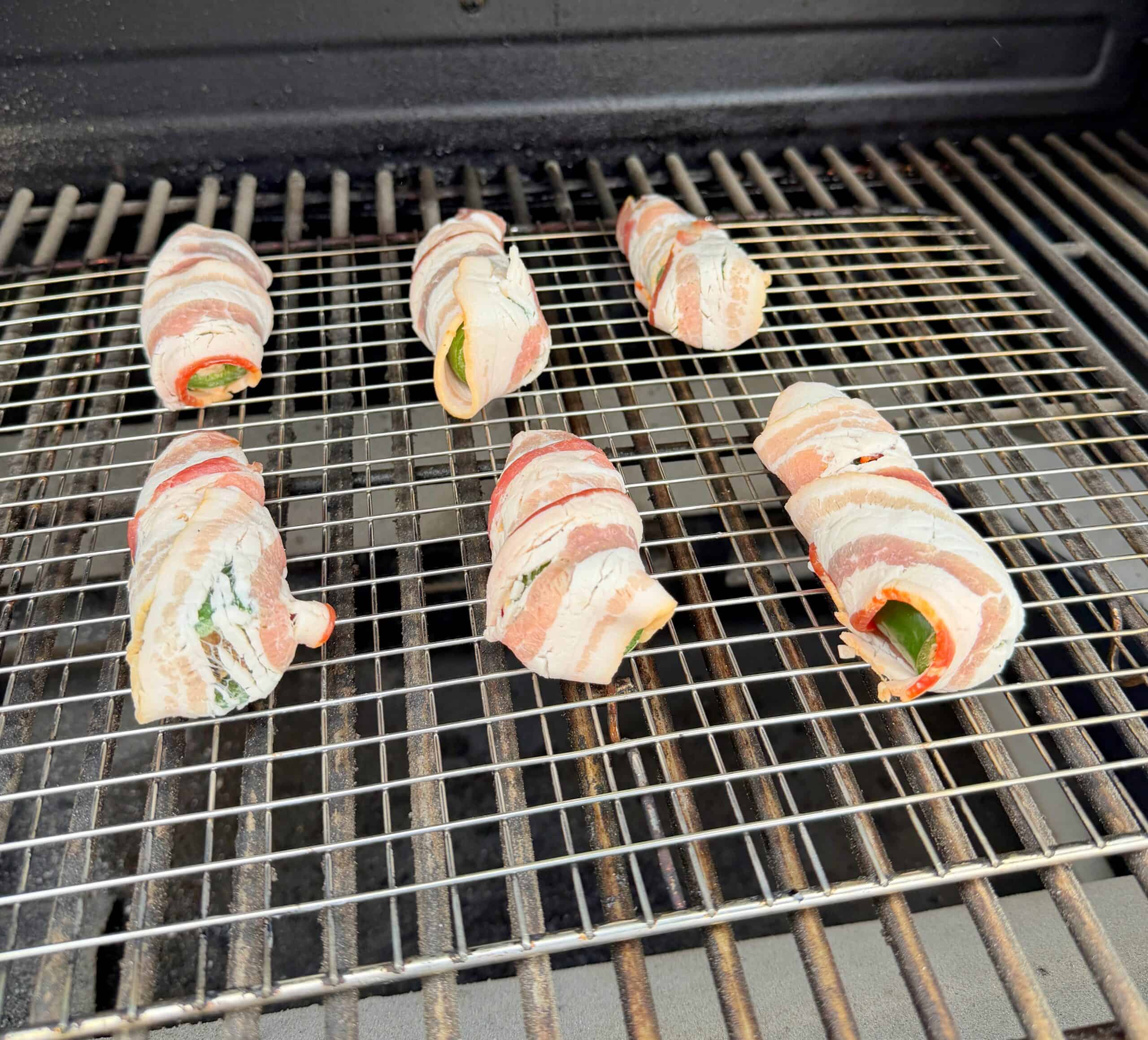Six bacon-wrapped jalapeño poppers are cooking on a grill, spaced apart on a metal grate. The jalapeños are stuffed and wrapped with raw bacon, ready to be grilled.