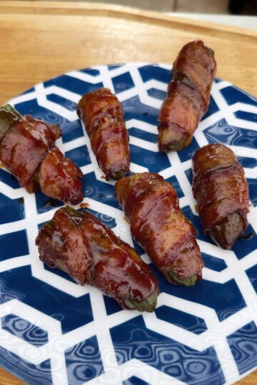 Six bacon-wrapped jalapeño poppers, known as Texas Twinkies, are glazed and roasted, then arranged on a white and blue geometric-patterned plate atop a wooden surface.