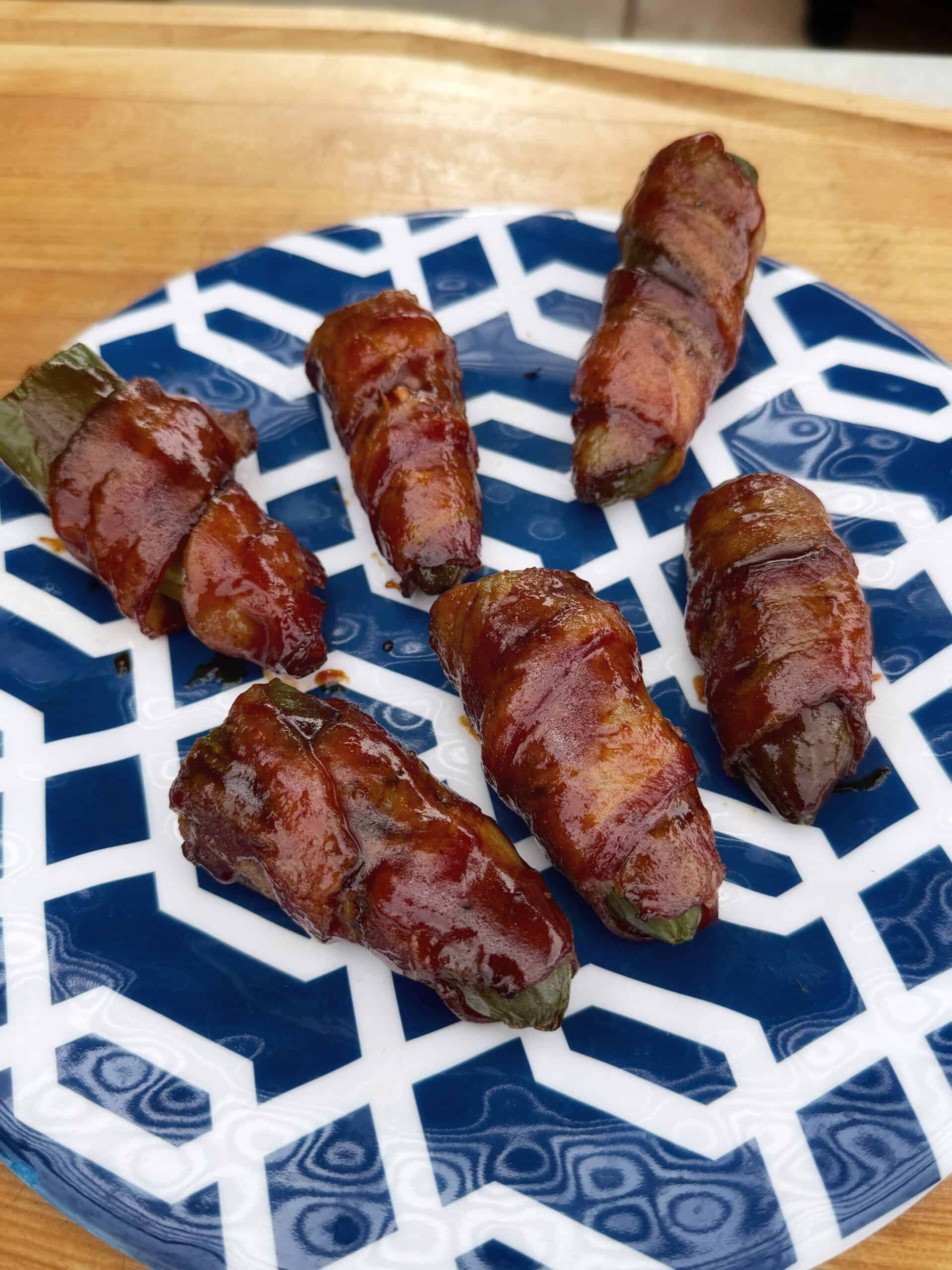 Six bacon-wrapped jalapeño poppers, known as Texas Twinkies, are glazed and roasted, then arranged on a white and blue geometric-patterned plate atop a wooden surface.