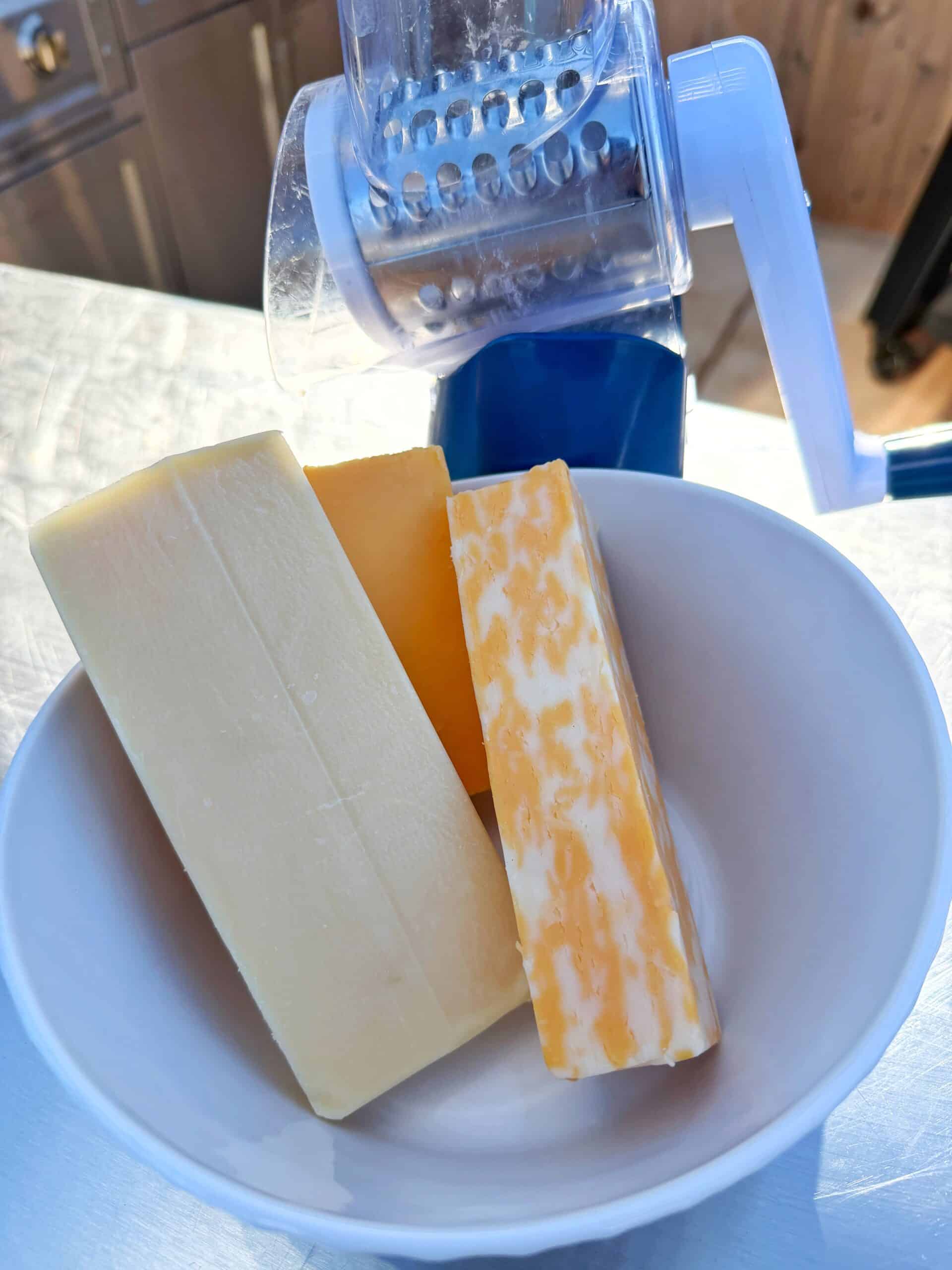 A white bowl contains three blocks of cheese—one white, one orange, and one marbled—sitting in front of a blue and white rotary cheese grater on a light-colored surface.