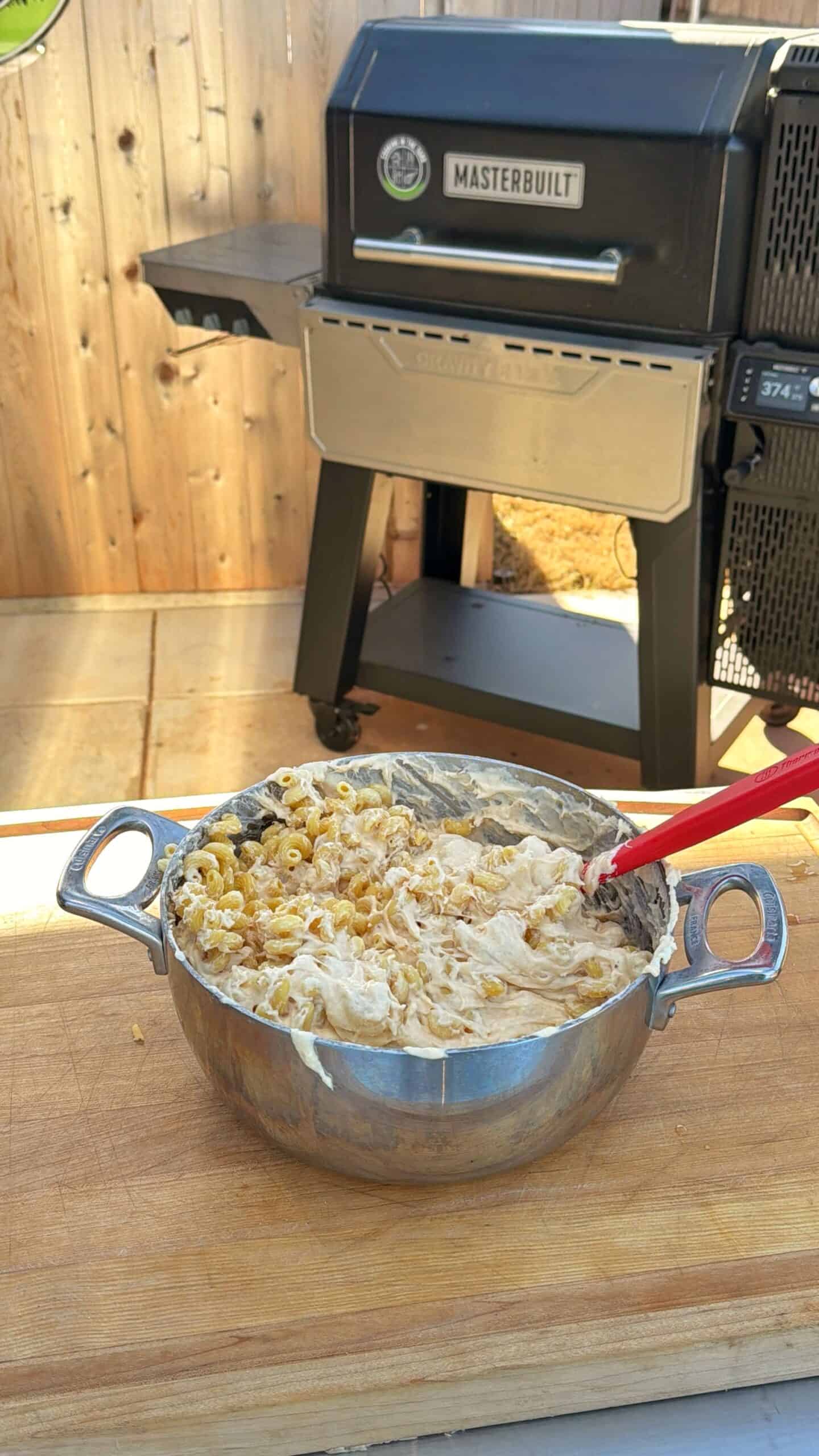 A large metal bowl of creamy pasta with shredded chicken is being mixed with a red spatula on a wooden surface outdoors, with a Masterbuilt smoker grill in the background.