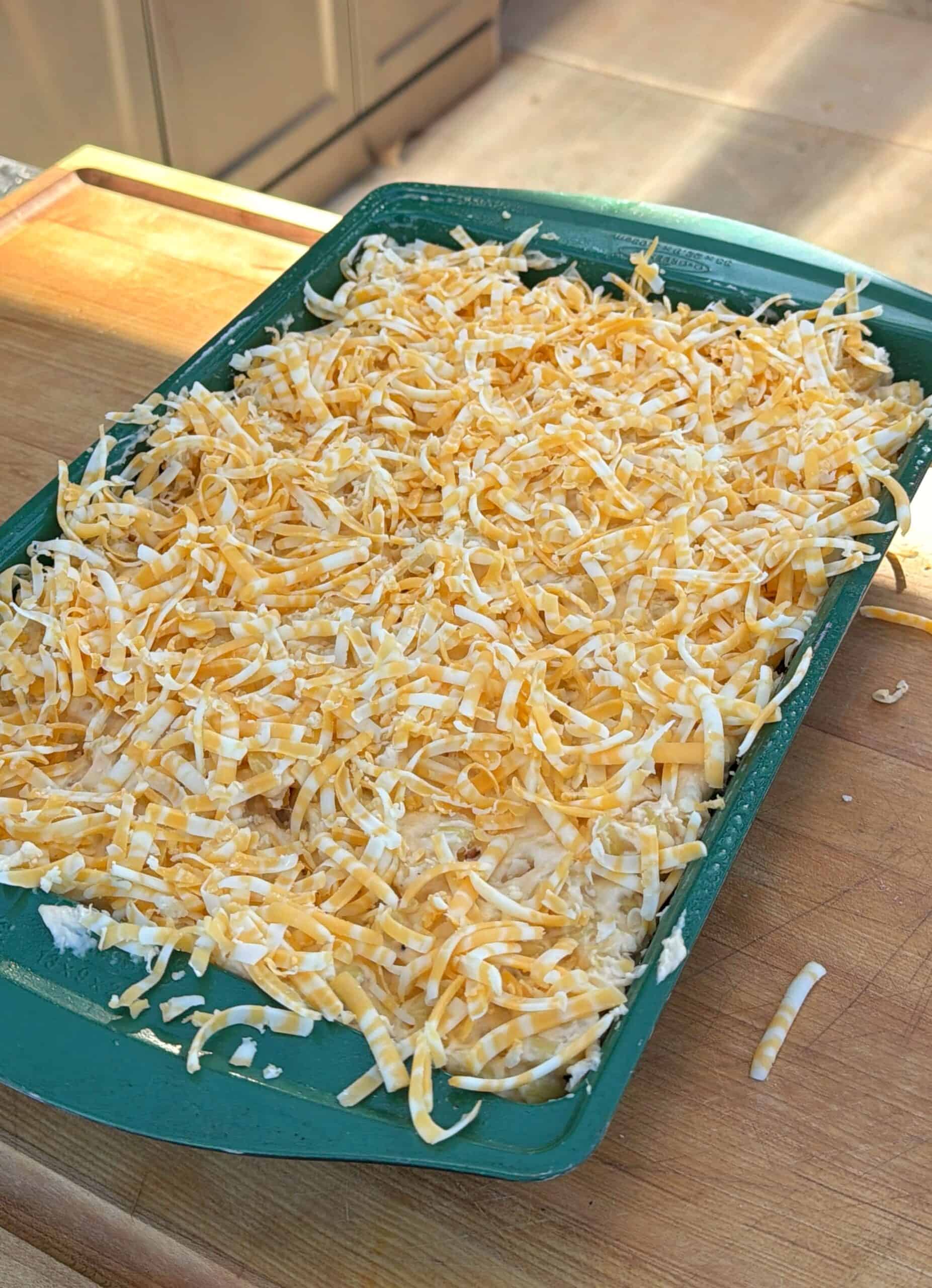 A green rectangular baking dish filled with a layer of shredded yellow and white cheese sits on a wooden surface, ready to be baked. Sunlight streams in from the side.