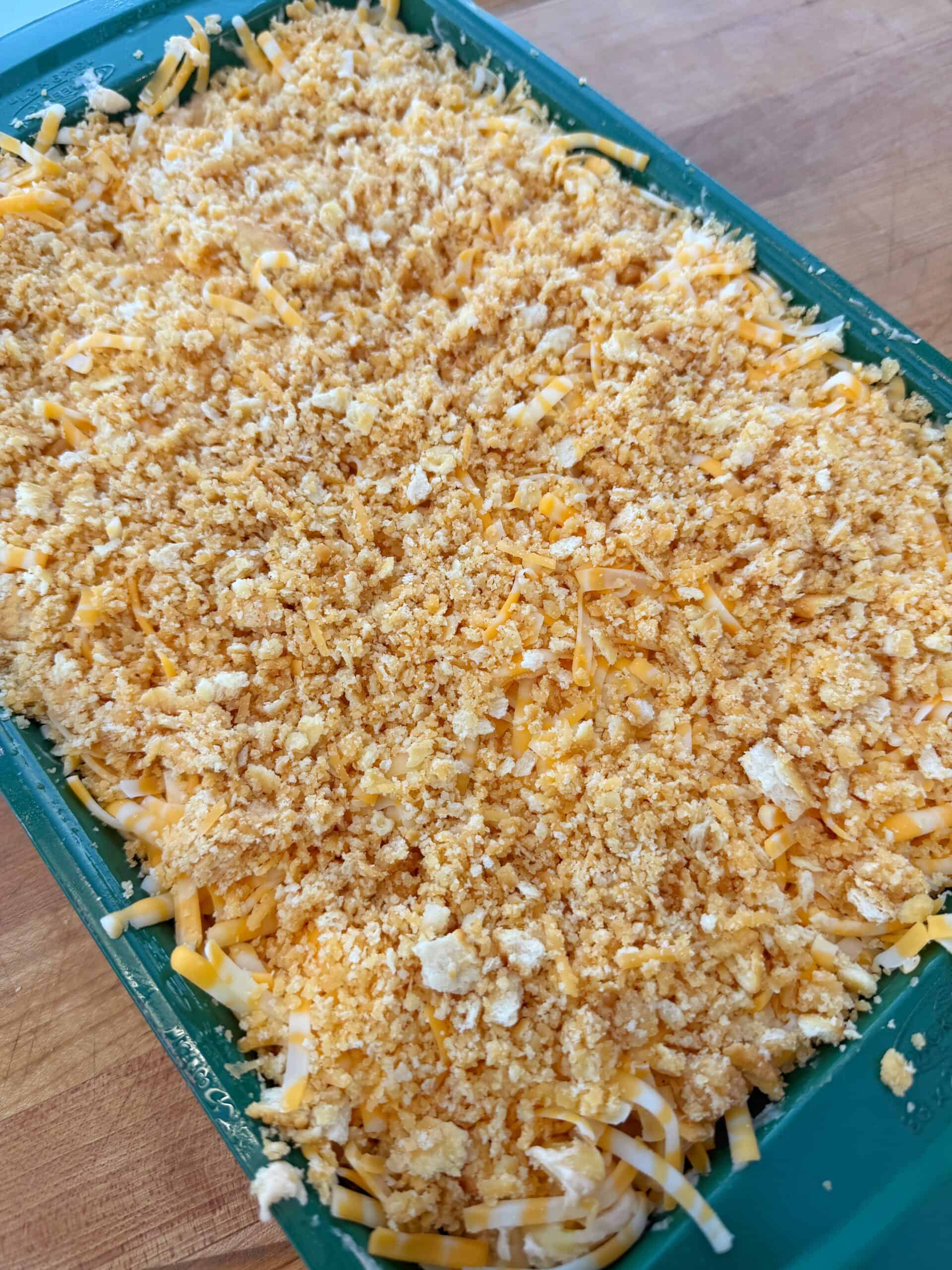 A close-up of an unbaked casserole in a green baking dish, topped with shredded cheese and a layer of crushed crackers on a wooden surface.
