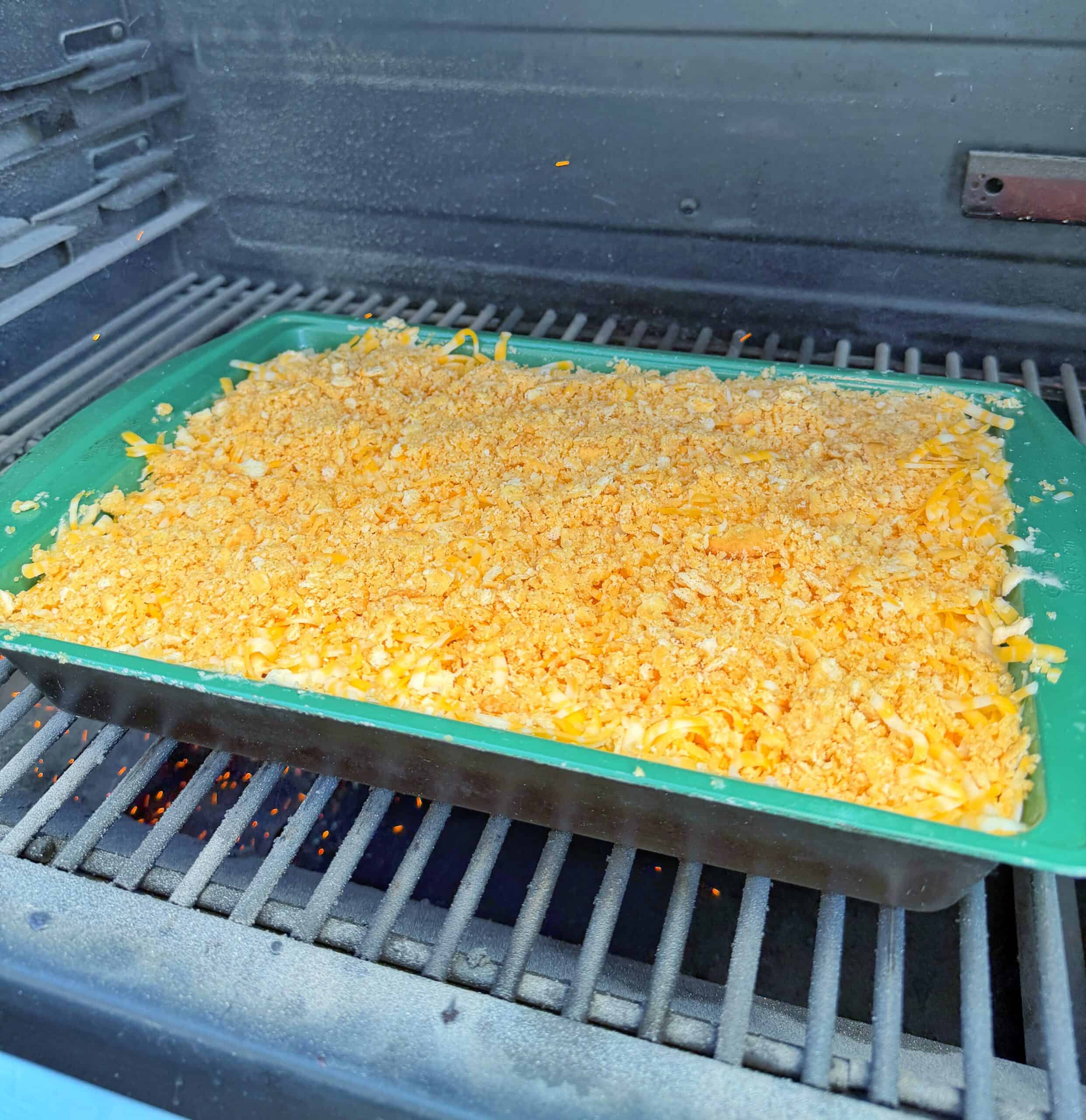 A green baking dish filled with macaroni and cheese topped with grated cheese and breadcrumbs sits on the grill rack inside a barbecue smoker.