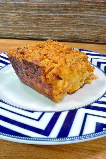 A slice of golden, crumb-topped casserole sits on a white plate with a blue geometric border, set on a wooden surface with a rustic wood background.