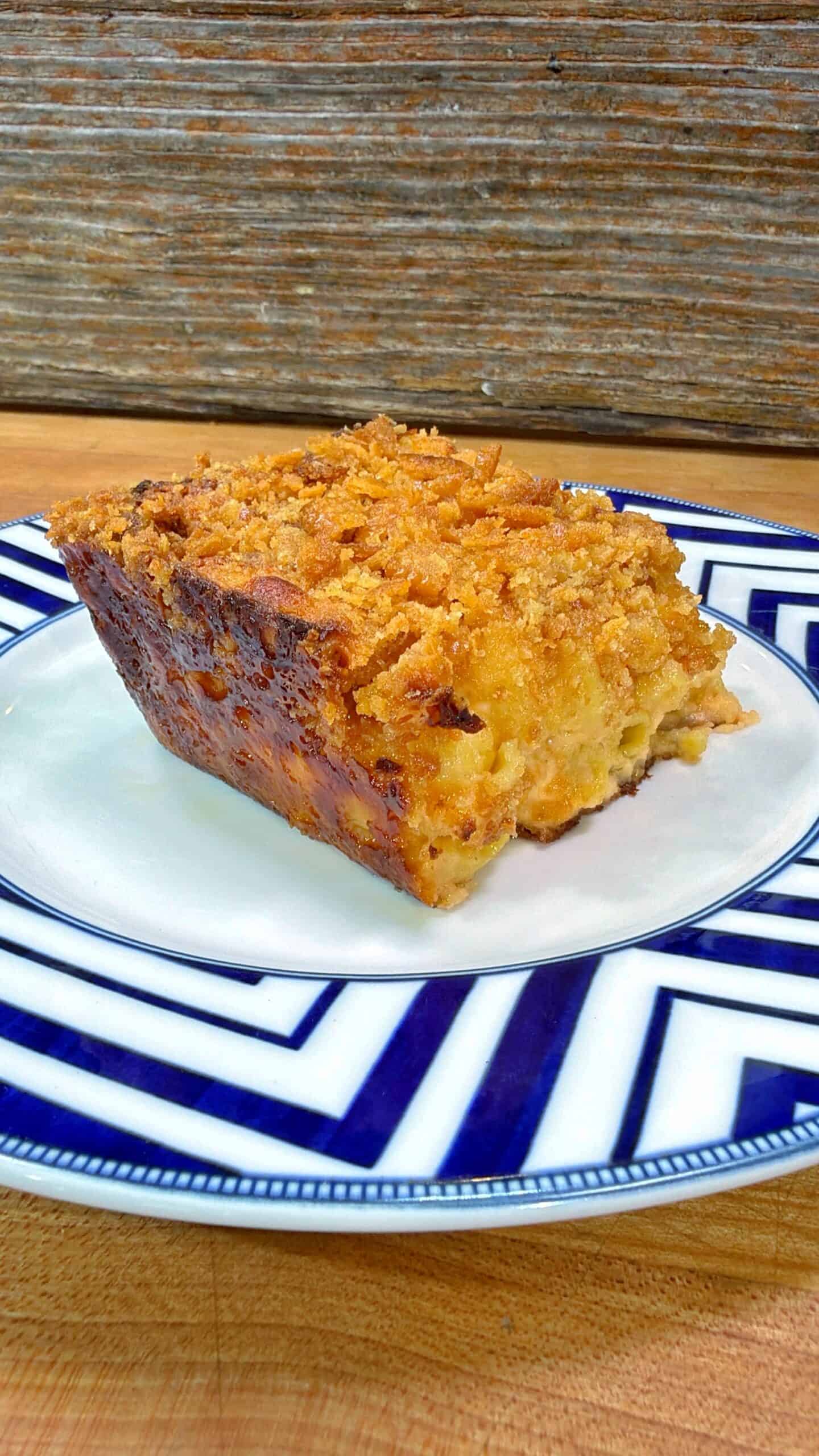 A slice of golden, crumb-topped casserole sits on a white plate with a blue geometric border, set on a wooden surface with a rustic wood background.