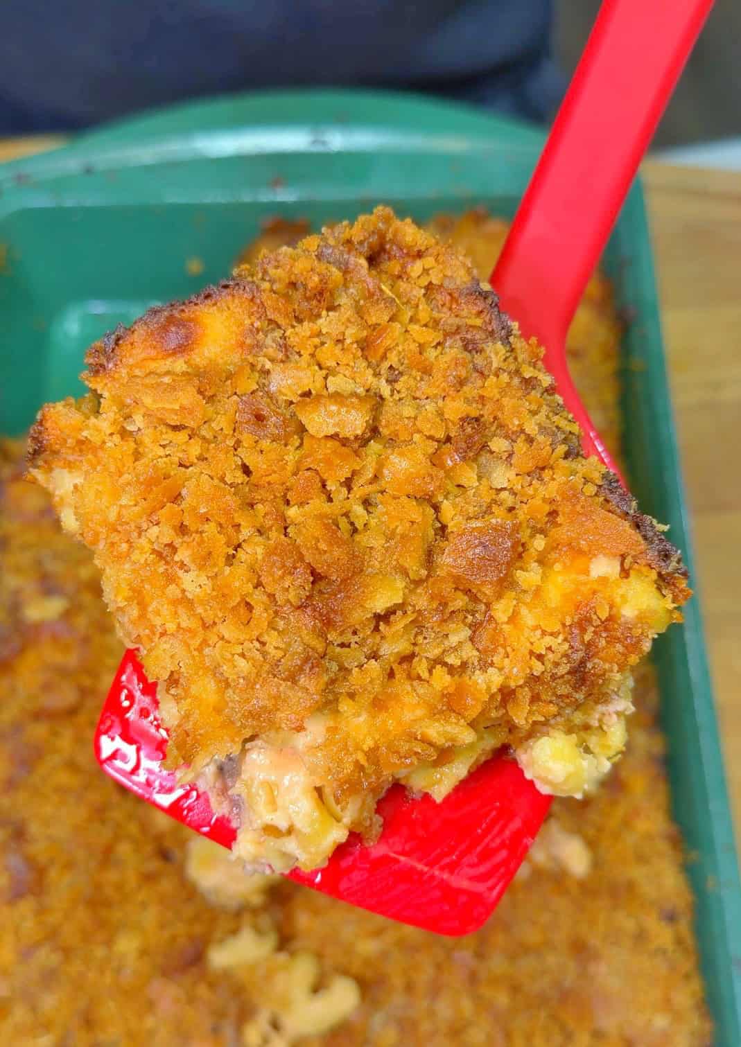 A close-up of a serving of smoked mac and cheese with a crispy, golden breadcrumb topping on a red spatula, held above a green baking dish.