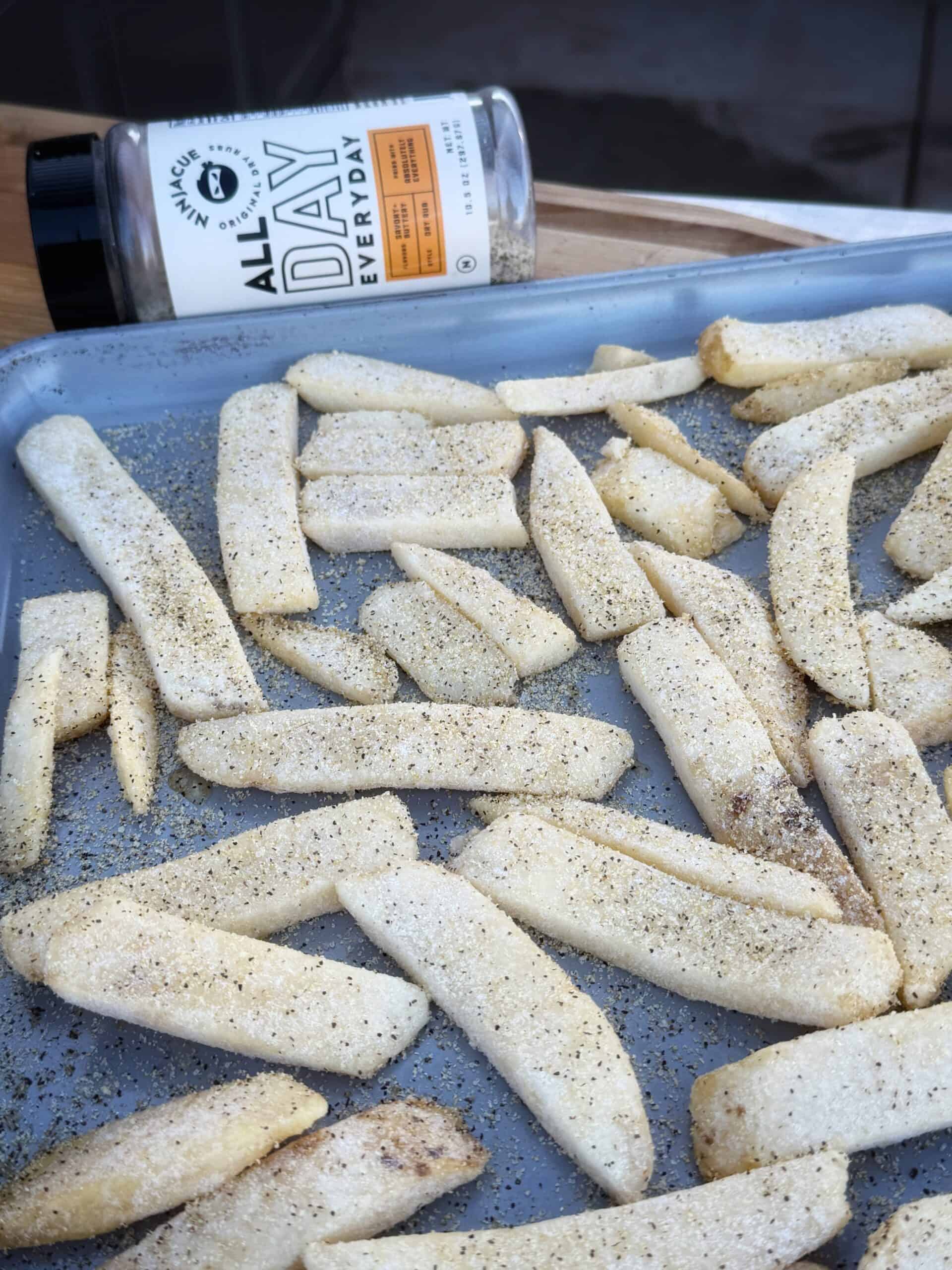 Frozen French fries spread on a baking sheet, sprinkled with seasoning. A container of All Day Every Day seasoning is lying on its side in the background.