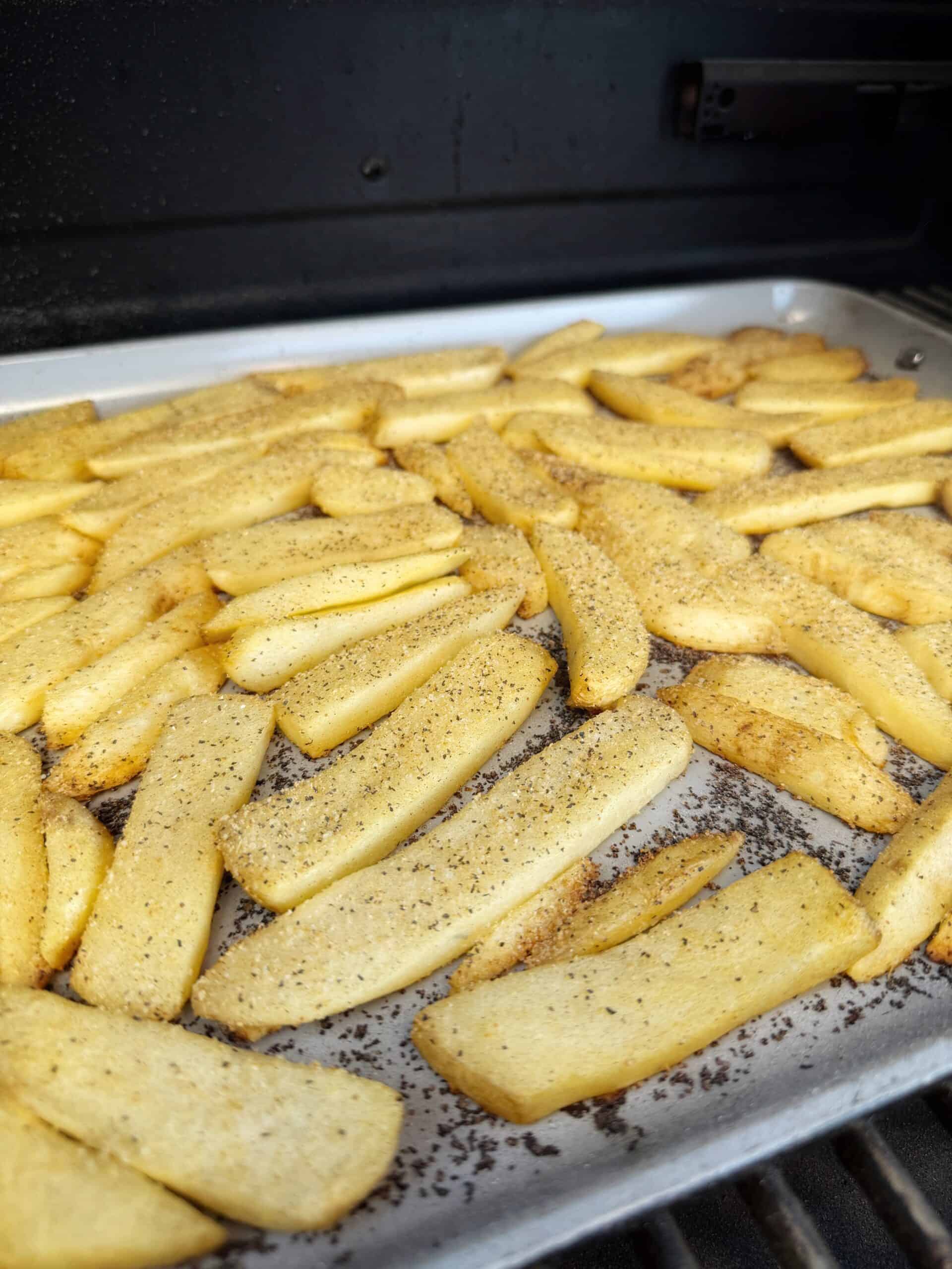 A baking tray filled with seasoned potato fries is placed inside an oven or grill, with the fries spread out evenly and lightly sprinkled with black pepper or spices.